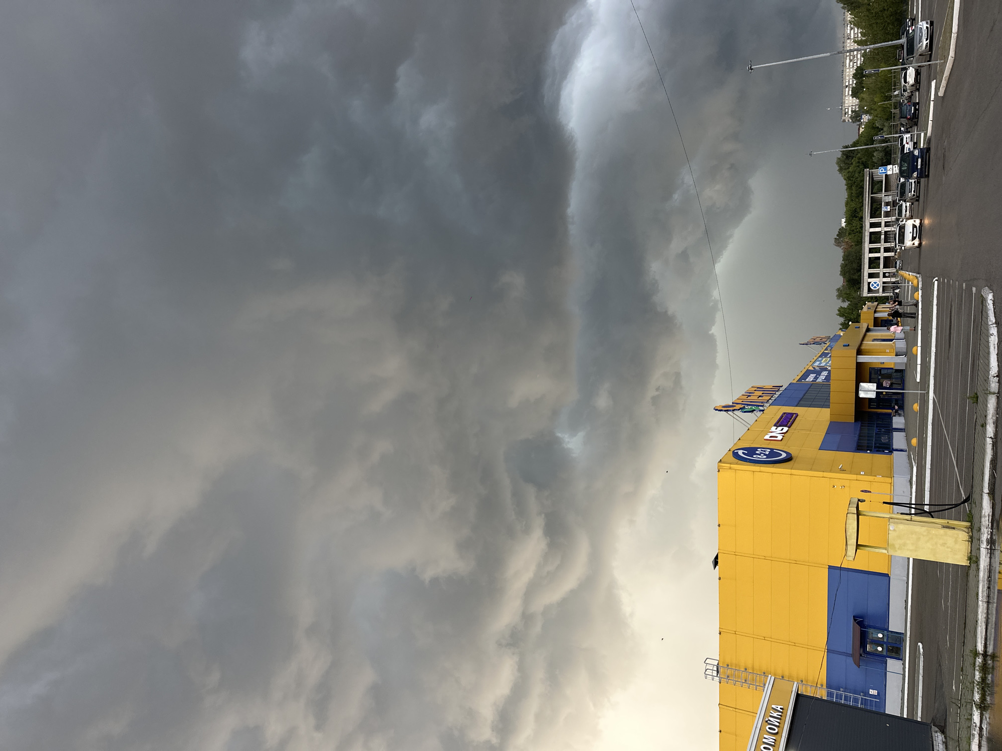 The image captures a stormy sky over a retail parking lot in Biysk, Russia. Dark, ominous storm clouds dominate the upper portion of the frame, casting a dramatic light on the scene below. In the midground, a bright yellow and blue supermarket, identified by signage as "ЛЕНТА" (Lenta) and "DNS", stands prominently. To the left of the supermarket, a car wash labeled "МОЙКА" (moyka) is visible. Several cars are parked in the lot, with some people standing near the supermarket entrance. A distinctive yellow, old-fashioned gas pump stands abandoned in the foreground, reflecting the muted light. In the background, trees and residential buildings can be seen, silhouetted against the turbulent sky. The overall impression is one of an approaching storm over a typical urban commercial area.