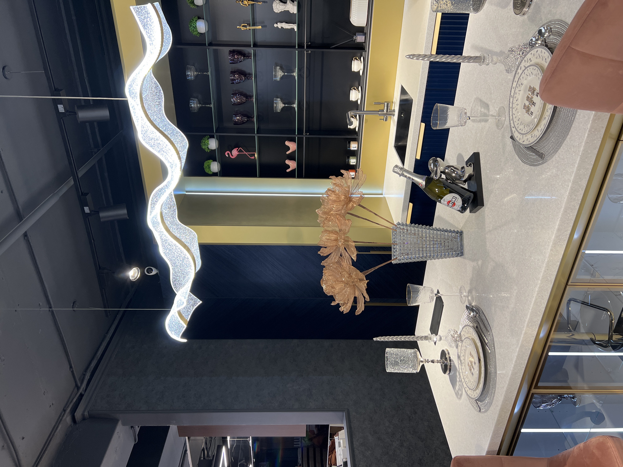 The image captures a contemporary interior, likely in a residential or commercial setting. It features a dining area with a white marble countertop table set for a formal meal. The table is adorned with plates, silverware, wine glasses, and decorative elements like a champagne bottle and a vase of artificial flowers. A modern, wavy chandelier hangs overhead, providing illumination.
In the background, there's a display cabinet with various decorative items arranged on shelves. The color palette includes black, gold, white, and shades of gray. The overall impression is that of a high-end, stylish space.
There is no definitive information to determine whether the image was taken in Moscow, Russia. The interior design style is common and can be found in many urban areas worldwide. Therefore, specifying Moscow as the location cannot be substantiated without further data.