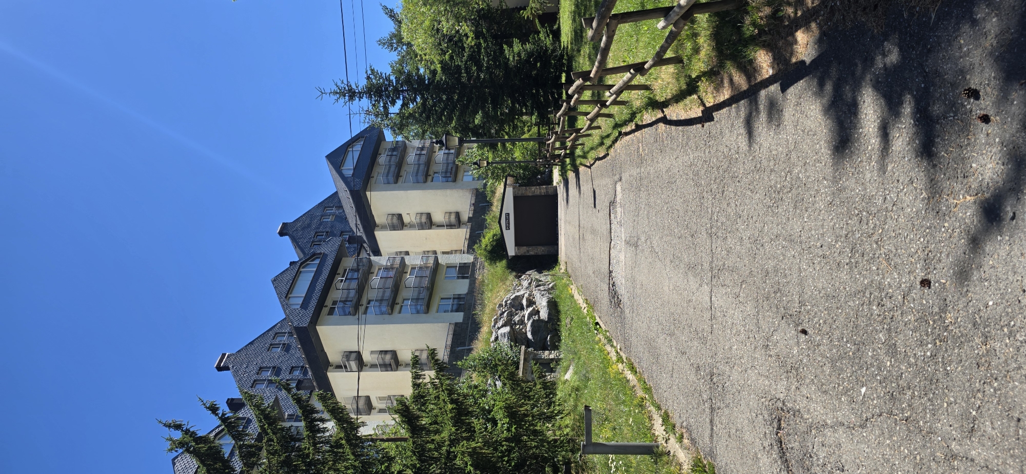 This media file is a photograph taken in Sallent de Gállego, Spain. The image features a multi-story building with a dark, angular roof, cream-colored walls, and balconies with metal railings. It appears to be a residential building or possibly a hotel, set against a clear blue sky. Trees and greenery surround the building, suggesting a natural, possibly mountainous, environment.
In the foreground, a paved pathway or road runs alongside a wooden fence. The path is textured with cracks and some debris, including pine cones and small dark objects, hinting at the natural surroundings. Shadows from trees are cast across the path, indicating a time of day when the sun is at an angle, likely mid-morning or afternoon. The weather appears to be sunny and pleasant.
To the right of the pathway, there is a small building with a dark garage door and a white facade. Text is visible above the garage door, appearing to read "PARKING". A rocky outcrop is also visible near the building, further reinforcing the impression of a natural, possibly mountainous, landscape. The overall scene conveys a sense of tranquility and a holiday or residential setting within a natural environment. No people are visible in the photograph.