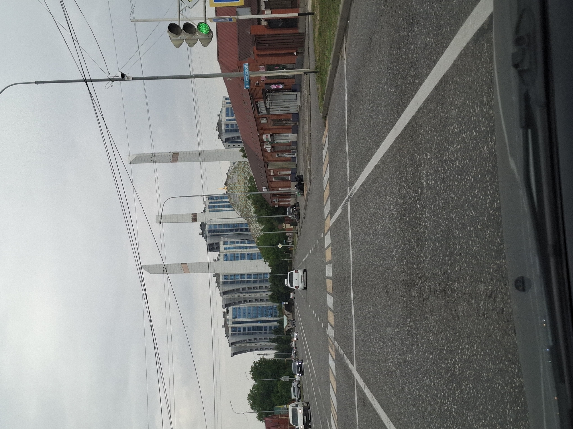 The image shows a street scene in Argun, Russia, with a pedestrian crossing in the foreground. In the background, there are modern apartment buildings with distinctive curved facades and white tower-like structures. A red brick building with a sign in Cyrillic script that reads "Н. КРУПСКОЙ" is visible on the right side of the street. A traffic light with a green signal is also present. Several cars are driving on the road, including a white van, a black sedan, and a white hatchback. The sky is overcast, and the overall atmosphere is that of a cloudy day.