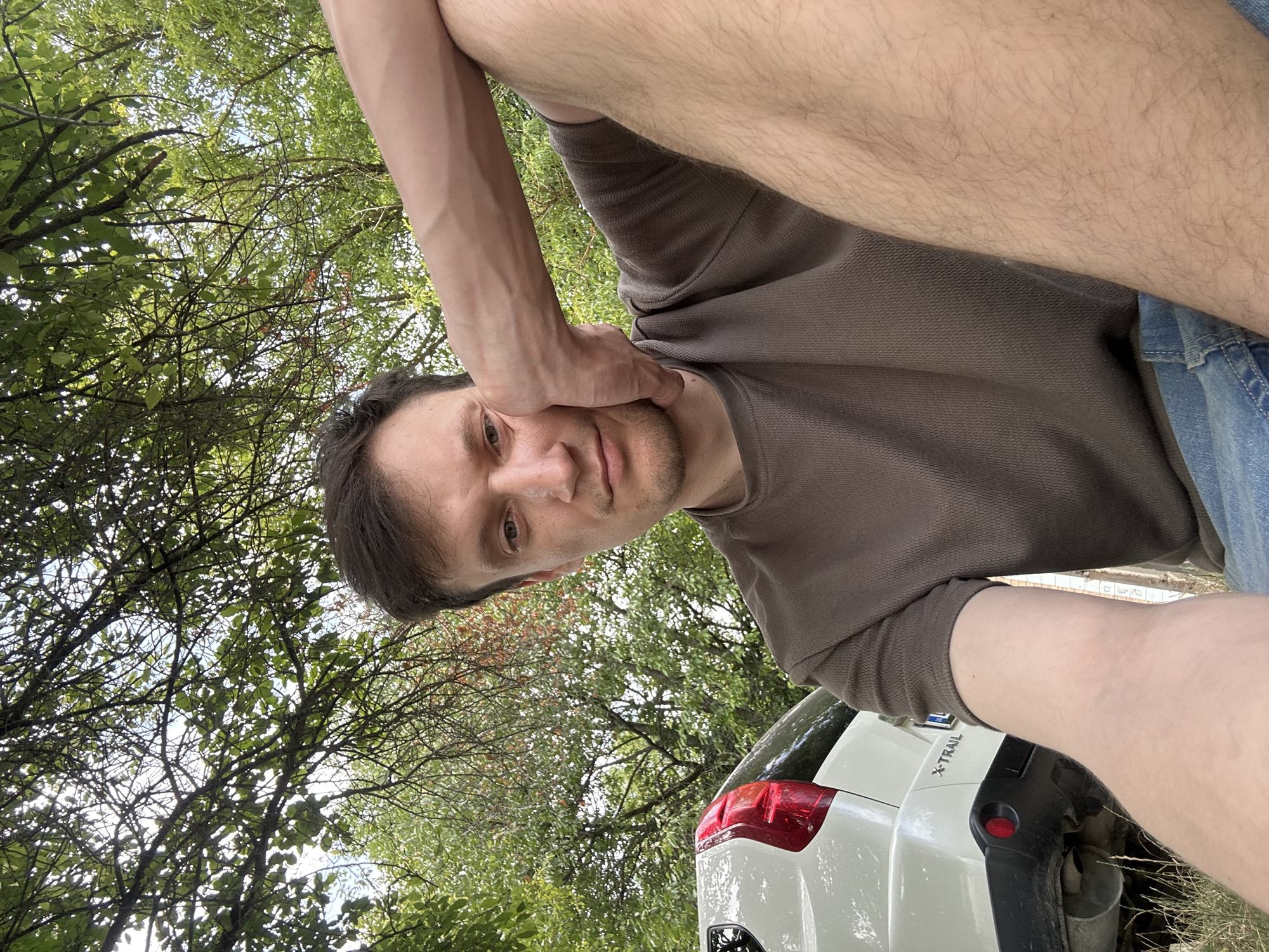 In Makiv, Ukraine, a man is pictured outdoors, seated in front of a white Nissan X-Trail SUV. He is wearing a brown t-shirt and blue jeans. The man is resting his head on his hand, with his arm bent at the elbow. His gaze is directed towards the camera. The background consists of lush green foliage and trees, with dappled sunlight filtering through the leaves. The overall scene suggests a casual, outdoor setting, possibly a park or a rural area.