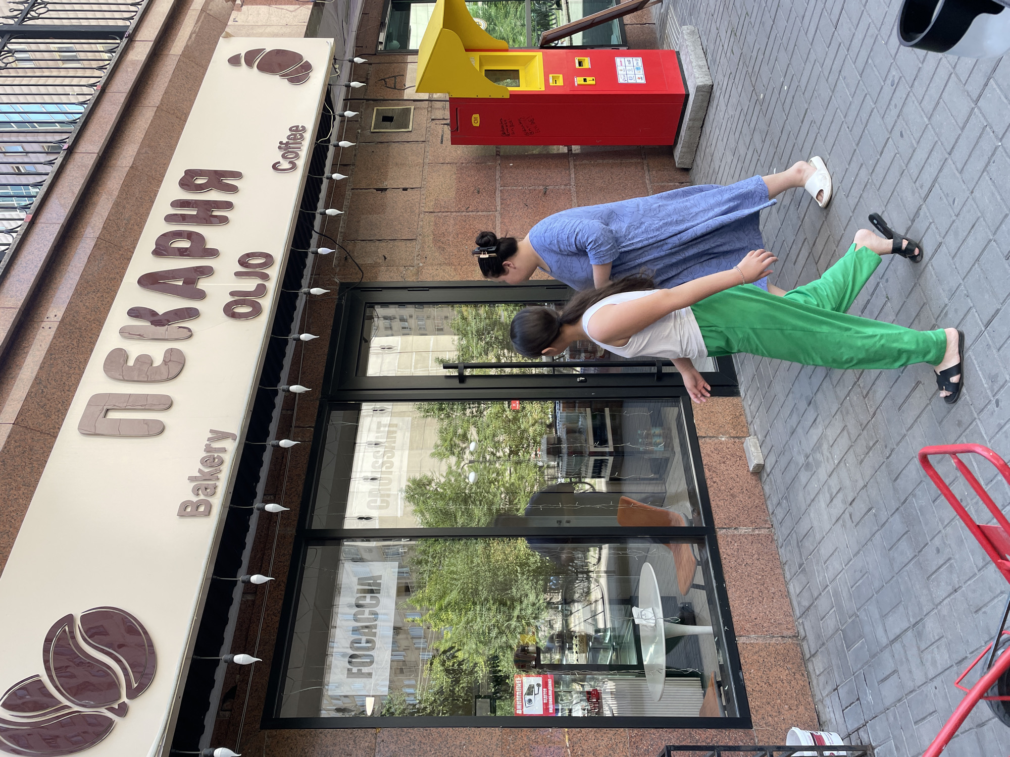 The image shows the exterior of a bakery in Almaty, Kazakhstan. The bakery's sign reads "ПЕКАРНЯ OLJO Bakery Coffee" with images of coffee beans and a croissant. The storefront features large glass windows displaying tables and chairs inside, along with signs for "FOCACCIA" and "CROISSANT." Two women are walking on the sidewalk in front of the bakery. One woman, on the right, is wearing a blue dress and white sandals, while the other, on the left, is wearing a white top and green pants, and black sandals. A red and yellow vending machine is visible to the right of the bakery entrance. The sidewalk is paved with gray tiles. The building facade is made of brown granite. A red handcart is partially visible in the foreground.