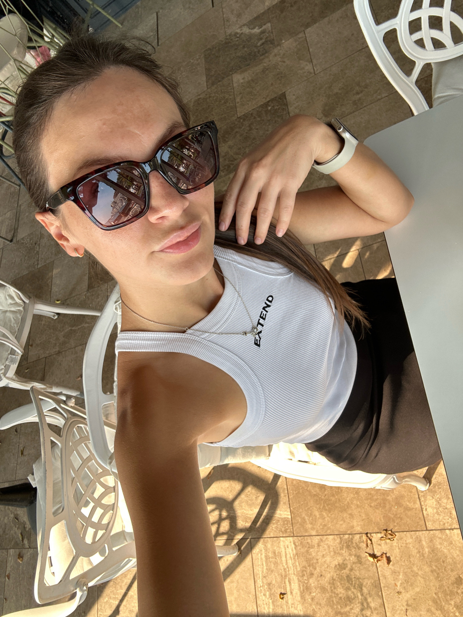 A high-angle, medium shot captures a woman from the chest up, taking a selfie outdoors. She is wearing large, tortoiseshell sunglasses and a white, ribbed tank top with the word "EXTEND" printed in black on the left side. A thin silver necklace with a small charm hangs around her neck. She is wearing a light-colored smartwatch on her left wrist. Her right arm is bent, with her hand resting on her head, and her left arm is bent at the elbow, resting on the table. She is sitting at a white table next to a white outdoor chair with an intricate woven design. The background shows a tiled patio bathed in sunlight, with shadows from the chair and table cast across it. The woman's hair is dark brown and pulled back from her face. The setting appears to be an outdoor seating area, potentially a cafe or restaurant in the city of Rafailovići, Montenegro, given the ambient light and outdoor furniture.