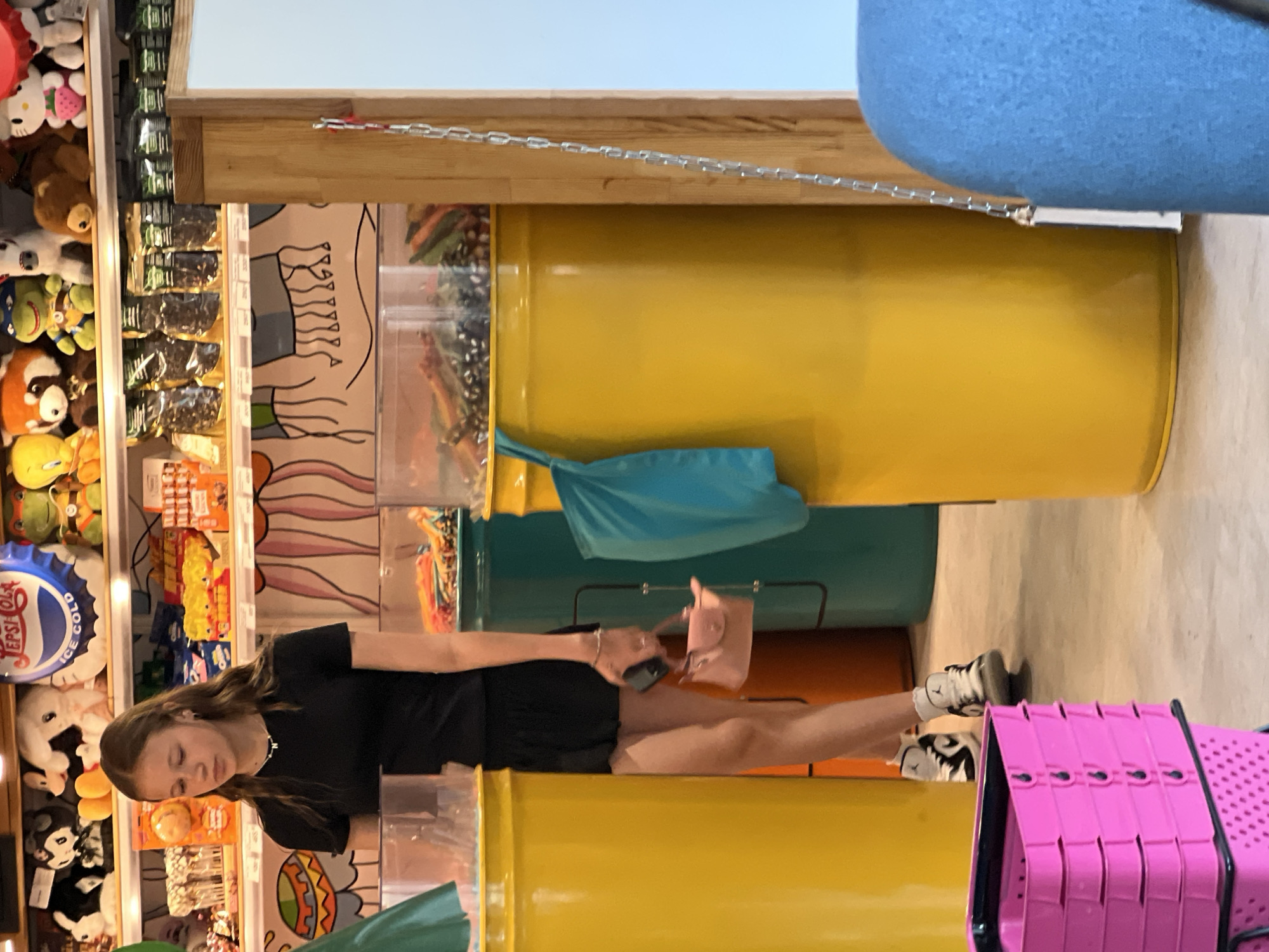 The image captures a young woman standing in a store filled with colorful displays, likely a candy or novelty shop. She is wearing a black t-shirt and black shorts, with white sneakers. In her left hand, she holds a small pink handbag. She is standing between two large yellow cylindrical bins, with a green bin behind her. The background shows shelves stocked with various products, including stuffed animals and packaged goods. A blue bag hangs on the right side of the frame. The floor appears to be concrete. The overall setting suggests a commercial establishment. The image does not provide any specific visual cues to indicate the location is in Novosibirsk, Russia.