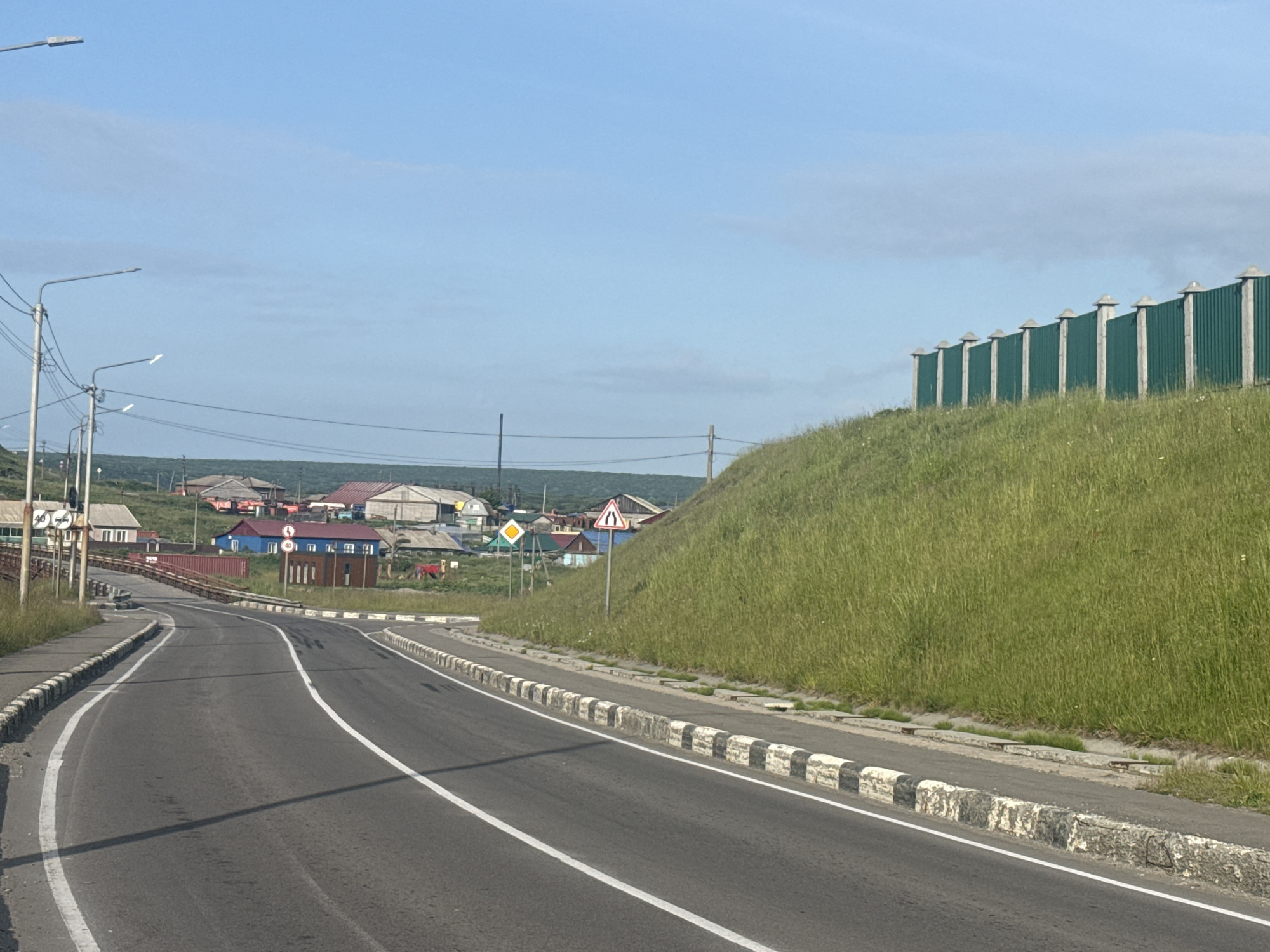 The image depicts a rural road leading into a small settlement. A paved road with white lane markings curves to the left and leads towards a cluster of houses. The houses are varied in color and construction, with some appearing to be made of wood. A green metal fence runs along a grassy embankment on the right side of the road. In the distance, a line of utility poles with streetlights is visible. The sky is a clear blue with a few scattered clouds. There are no people or vehicles visible in the scene. The overall impression is one of a peaceful, undeveloped area, possibly in a northern climate given the style of the houses and the lush green vegetation. The location is identified as Kuril'sk, Russia.
