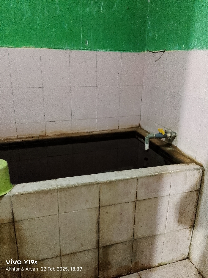The media file displays an indoor utility area or simple bathroom, likely in Kroya, Indonesia, characterized by a large, rectangular water basin. No people are visible in the image.
The central object is a waist-high, tiled basin filled with dark water, extending along two walls of the corner. The walls above the basin are tiled with off-white square tiles up to about mid-height, with the upper portion painted a vibrant green. A simple PVC pipe faucet with a yellow handle is attached to the right wall, directly over the water basin. To the left, a bright green plastic bucket or small basin rests on the edge of the large water container.
Notable details include the condition of the surfaces: the white tiles and grout lines around the water basin show significant staining and dirt accumulation, suggesting heavy use or a lack of recent cleaning. The water in the basin appears very dark, possibly due to depth, poor lighting, or murkiness. The overall lighting in the room is dim.
There is no activity currently taking place; the scene is static, depicting a facility. The setting is typical of traditional Indonesian bathrooms where such "bak mandi" (water reservoirs for bathing) are common.
Visible text in a watermark at the bottom left corner reads "vivo Y19s", "Akhtar & Arvan", and "22 Feb 2025, 18.39", indicating the device, potentially the photographers, and the date and time the photo was taken.
