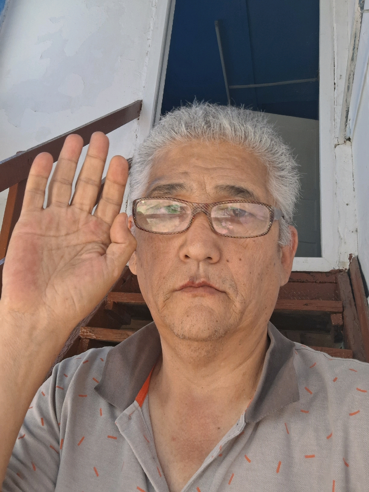 In the city of Almaty, Kazakhstan, a middle-aged East Asian man with grey hair and glasses is captured in a selfie. He is raising his left hand in a gesture that could be interpreted as a wave or a stop sign, with his palm facing the camera. The man is wearing a grey collared shirt with a subtle orange-and-dash pattern. He is positioned on wooden stairs, suggesting an outdoor or semi-outdoor environment, possibly a porch or entryway. The background includes white painted walls and a dark blue framed structure, possibly a window or door. The lighting indicates it is daytime.