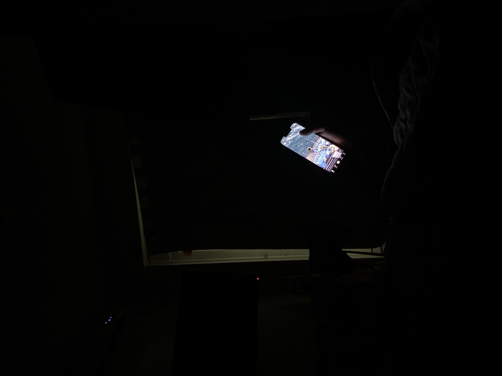 The image shows a person in a dark room holding a lit smartphone in their hand. A monitor or large screen is visible behind the phone. The rest of the environment is indistinct due to the darkness. There is no evidence in the image to indicate the location of the photo in Buenos Aires, Argentina, or any other city.