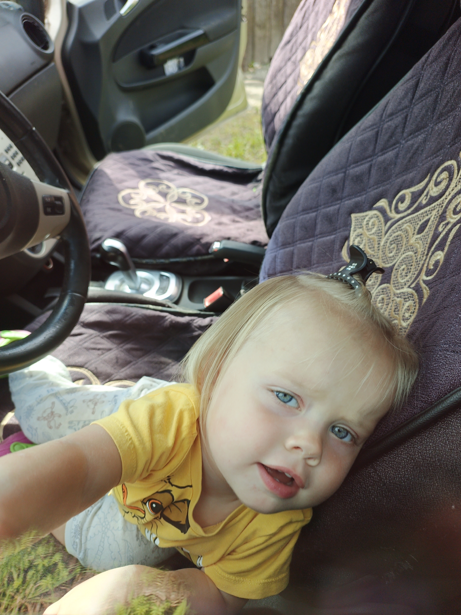 A young child with blonde hair and blue eyes is lying on the seat of a car. The child is wearing a yellow t-shirt with a cartoon character on it. The car's interior is visible, including the steering wheel, gear shifter, and car door. The seat covers are dark purple with an embroidered design. The scene appears to be outdoors, with green grass visible in the background. This photograph was taken in the city of Medvedyovka, Russia.