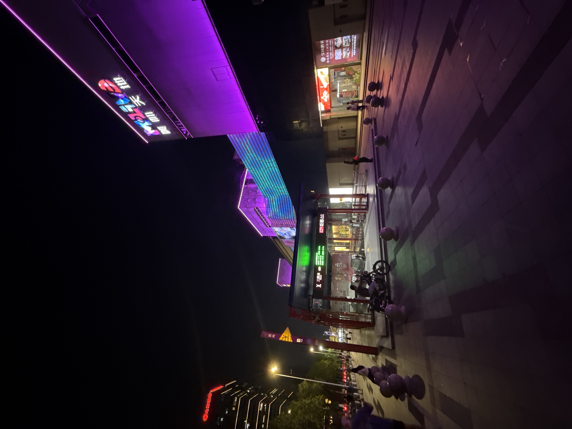 A nighttime, wide-angle shot captures a vibrant urban scene in Tianhe District, China, dominated by illuminated commercial buildings and pedestrian activity. In the upper right, a building facade is bathed in purple light, featuring a large, multi-colored neon sign that appears to read "mola" followed by Chinese characters. Another building to the left displays a horizontal band of green and blue illuminated panels, also part of the building's facade.
In the foreground, a paved walkway is visible, with several spherical bollards positioned on the left side. To the right, a covered structure, possibly a bus stop or subway entrance, is illuminated. Inside, escalators are visible, and people are present. A person is seated on a motorcycle parked in front of this structure.
Further to the left, a tall, modern building with numerous illuminated windows rises into the night sky. Streetlights and other ambient lighting contribute to the overall illumination of the scene. Several individuals are walking on the sidewalk and street, some silhouetted against the light. The atmosphere is lively, indicative of a commercial and entertainment area at night. The ground is paved with large tiles, reflecting the colorful lights from the buildings.