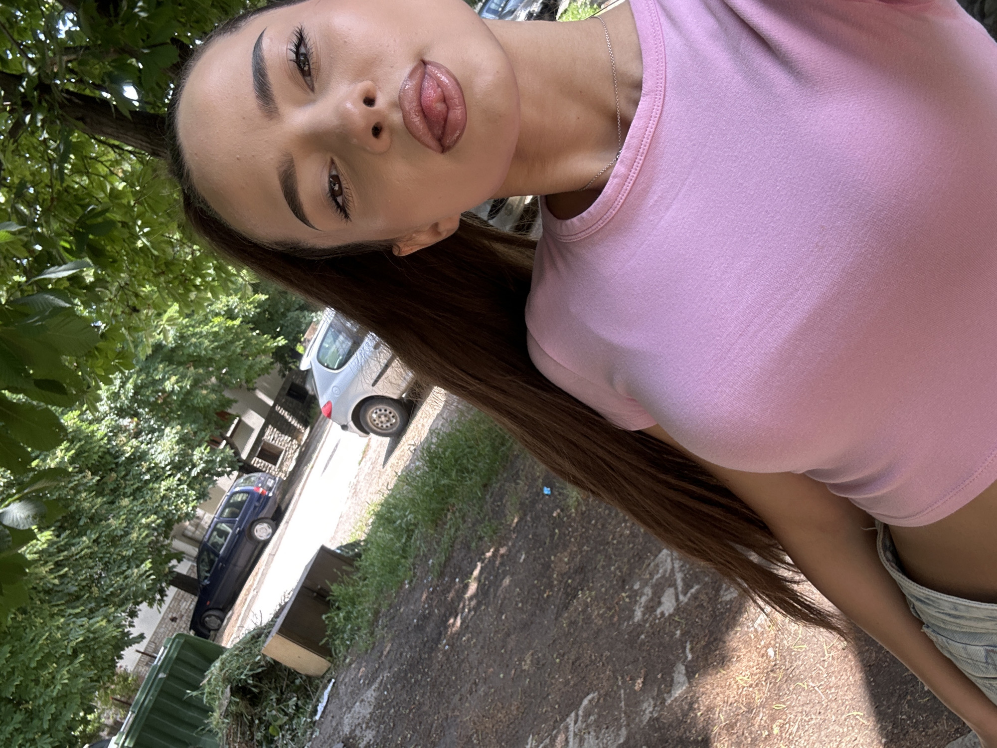 The image is a tilted, close-up selfie of a young woman taken outdoors. She is the primary subject, with long, straight dark brown hair, dark eyebrows, and defined eyelashes. She wears a light pink, short-sleeved crop top and light-wash denim shorts or jeans, along with a thin silver necklace. Her head is tilted, and she poses with her lips slightly parted, revealing the tip of her tongue in a playful expression.
The setting is an outdoor, possibly urban or suburban environment, bathed in bright daylight, indicating a sunny day with clear skies. Lush green foliage from trees dominates parts of the background. A street or paved area runs behind her, where a silver car and a dark blue car are parked. Green trash bins are also visible on the side of the path, which consists of dirt and patchy grass.
No visible text is present in the image.