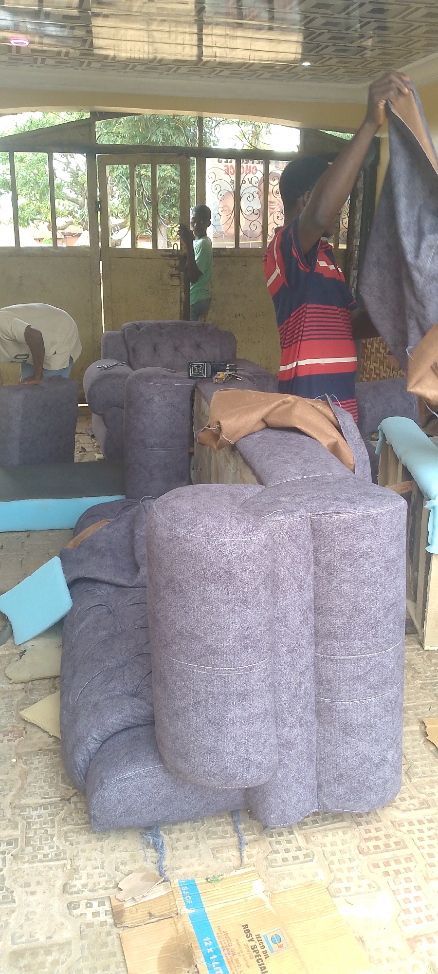 In an outdoor workshop, possibly in a developing urban area, the construction or upholstery of furniture is underway. The foreground is dominated by a partially assembled plush purple sofa, its tufted cushions and rolled armrests indicating a Chesterfield-style design. Several pieces of light blue foam padding are scattered around the sofa and a dark gray ottoman. In the middle ground, a man in a red and blue striped t-shirt is holding up a piece of dark fabric, appearing to be examining it or preparing to attach it. To his left, a young boy in a light green t-shirt stands near a metal gate. Behind the gate, lush green trees are visible, and a faded poster with an image of a person's face and text in a foreign language is affixed to the wall. In the background, another person, mostly obscured, is bent over, possibly working on another piece of furniture. The ground is a rough, uneven surface covered in light-colored tiles and debris, including pieces of cardboard. A cardboard box with text in a foreign language, including "JEZCO OIL," is prominently displayed on the ground in the lower right portion of the image. The lighting suggests it is daytime, with sunlight filtering through an overhead structure, creating shadows.