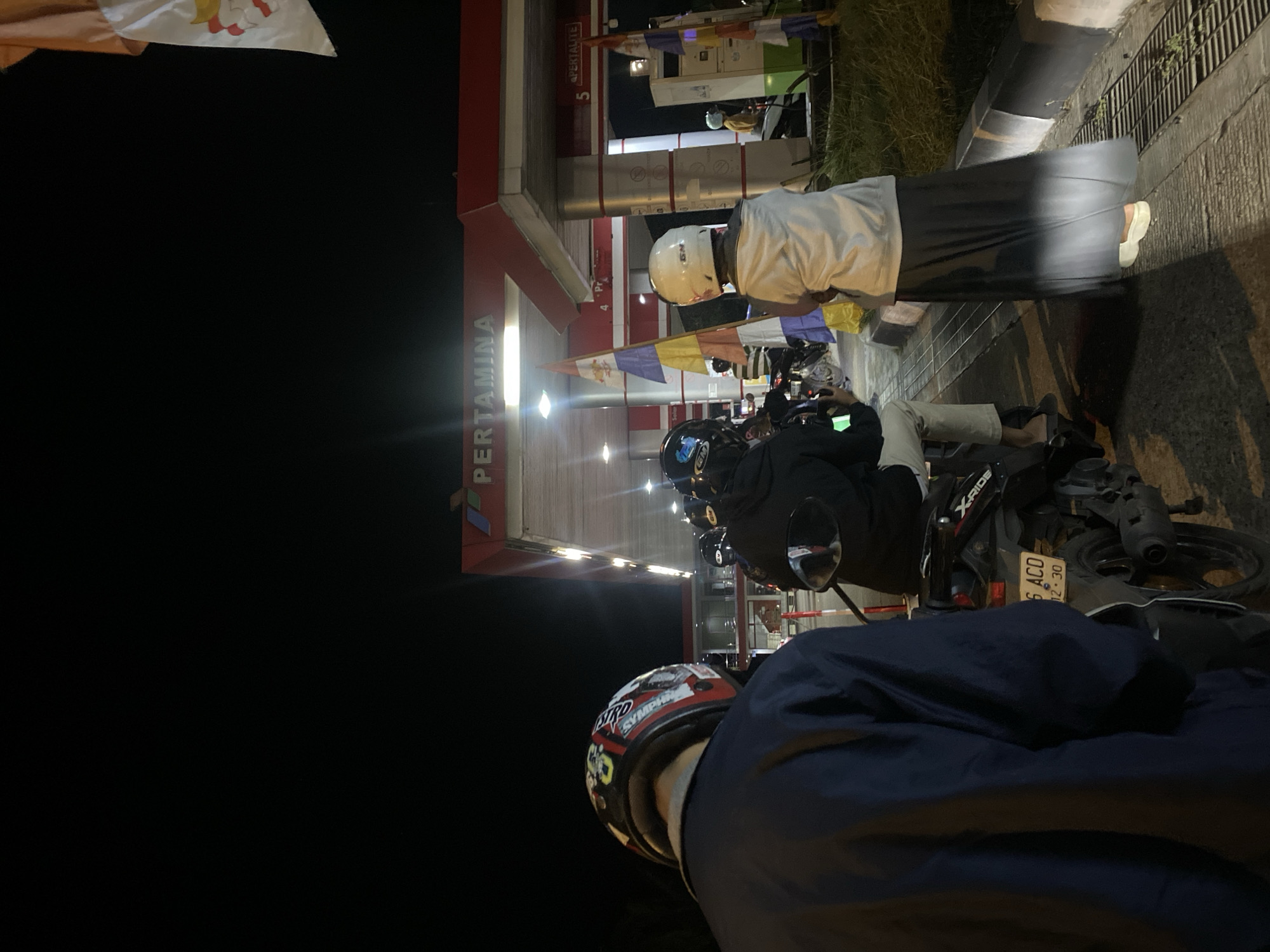 The image shows a nighttime scene at a Pertamina gas station in Gambut Barat, Indonesia. Several people on motorcycles are present, seemingly waiting in line. The gas station features prominent red and white branding, with illuminated pumps and a canopy. Flags with colorful panels are visible, attached to poles. The foreground is dominated by the back of a person wearing a dark blue jacket and a patterned helmet, facing away from the camera. To their right, another person in a white helmet and a light-colored hooded jacket with a long dark skirt is also visible. The ground is paved, with a drainage grate to the right. The overall atmosphere is that of a typical urban refueling stop at night.