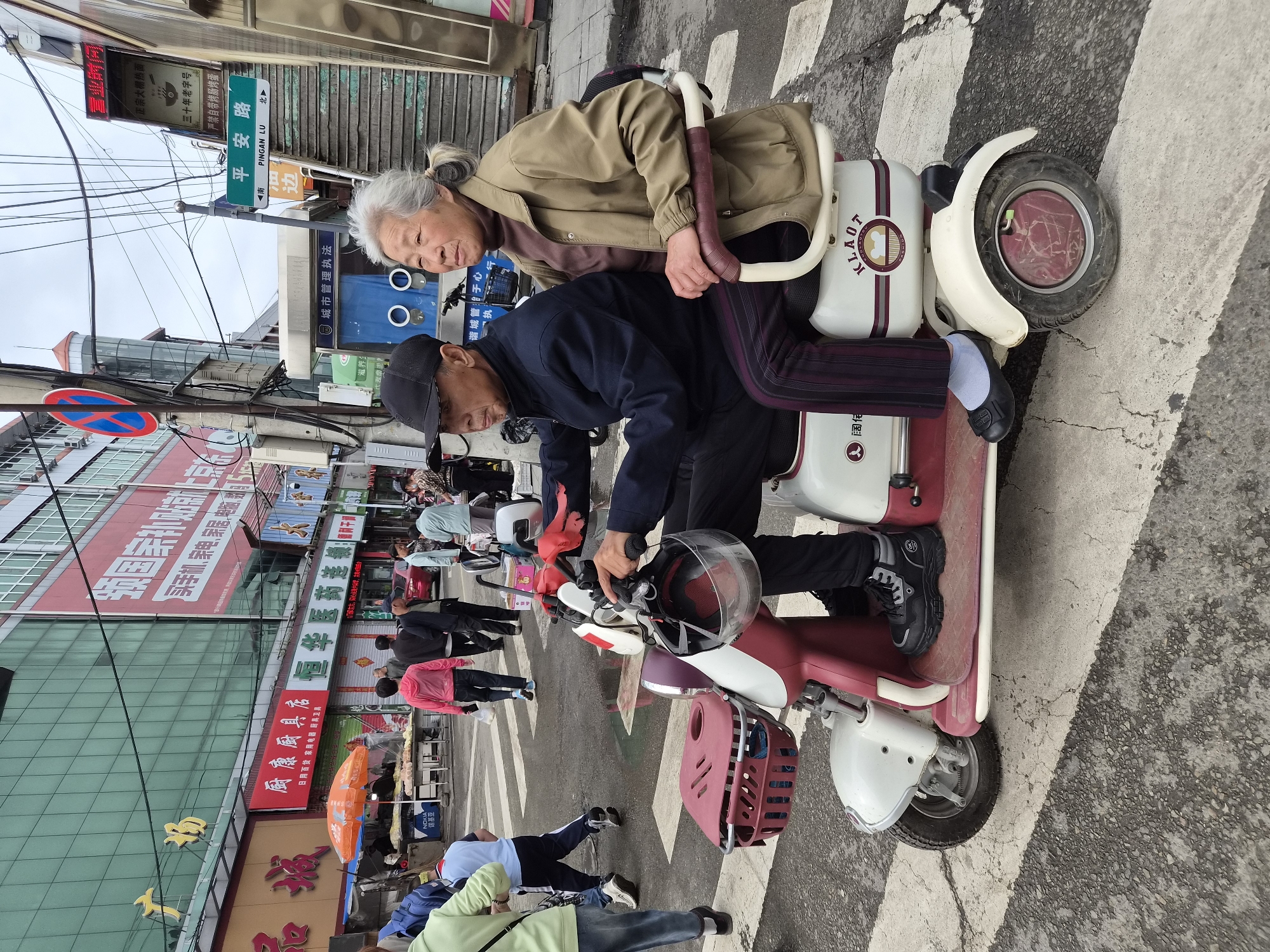 In Mishan, China, an elderly couple rides an electric scooter across a crosswalk. The man, wearing a black baseball cap and a dark jacket, drives the scooter. The woman, with gray hair pulled back in a ponytail, sits behind him, wearing a tan jacket over a brown turtleneck. The scooter has a red basket on the front and is decorated with a logo that appears to read "KLAOT". In the background, a bustling street scene unfolds with pedestrians, storefronts, and overhead electrical wires. The sky is overcast.