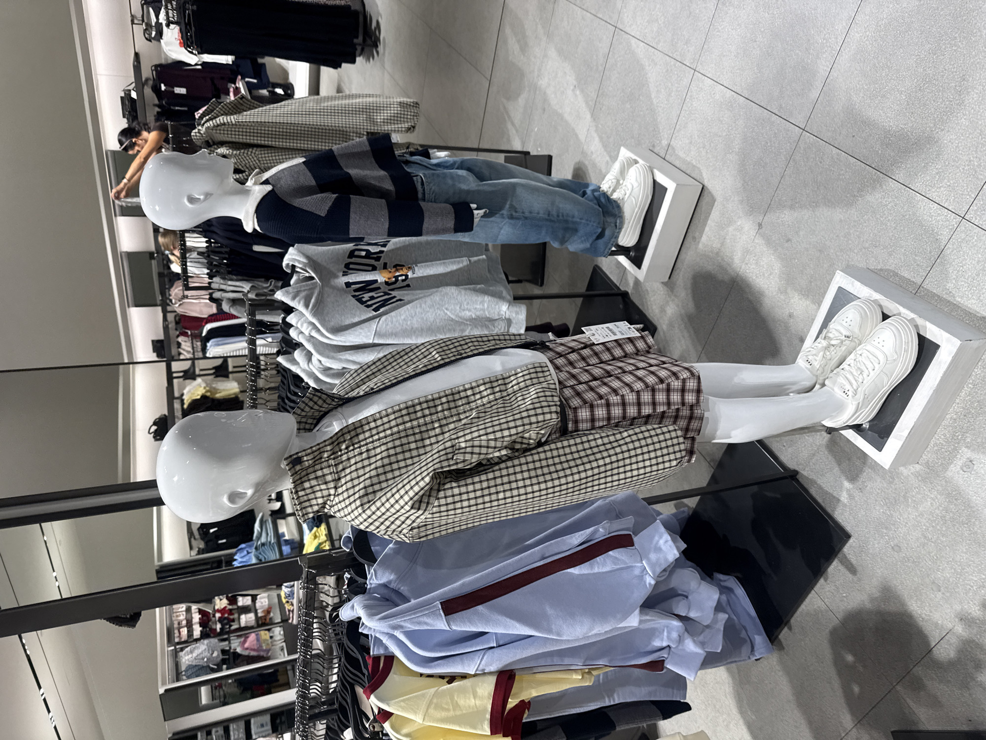The image depicts the interior of a clothing store in Tallinn, Estonia, likely at the Zara store in the Kristiine Keskus shopping mall, given the layout and merchandise. Two child mannequins are prominently displayed, showcasing casual wear. The mannequin on the left wears a plaid jacket over a white shirt and a plaid skirt, paired with white sneakers. The mannequin on the right is dressed in a striped long-sleeved shirt, blue jeans, and white sneakers. In the background, racks of clothing are visible, including t-shirts, hoodies, and jackets, organized on metal rails. The store features a clean, modern design with light-colored tiled flooring and reflective ceiling panels. The lighting is bright and even, characteristic of a retail environment. No people are directly interacting with the mannequins or the merchandise in the foreground.