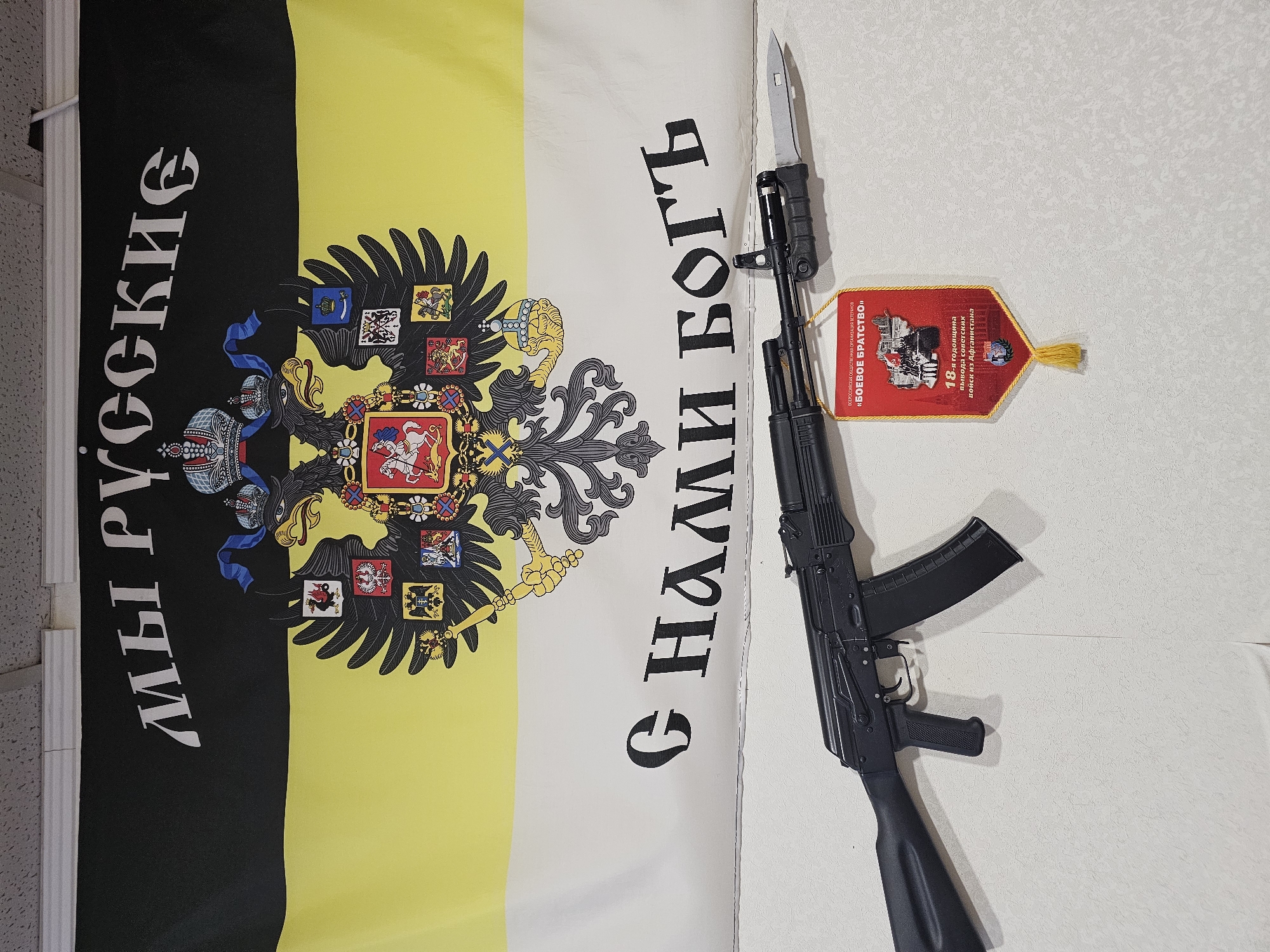 The image shows a black, yellow, and white flag with a black double-headed eagle emblem at the center. Text in Cyrillic script runs vertically along the sides of the flag: "МЫ РУССКИЕ" (We are Russians) on the left and "С НАМИ БОГЪ" (God is with us) on the right. A black AK-pattern assault rifle with a bayonet attached is laid over the flag. A red pennant with the inscription "БОЕВОЕ БРАТСТВО" (Combat Brotherhood) and the 18th anniversary of the withdrawal of Soviet troops from Afghanistan is near the gun. The photograph appears to be taken indoors against a wall, possibly in Russia. There is no mention of the city of Moscow, Russia.