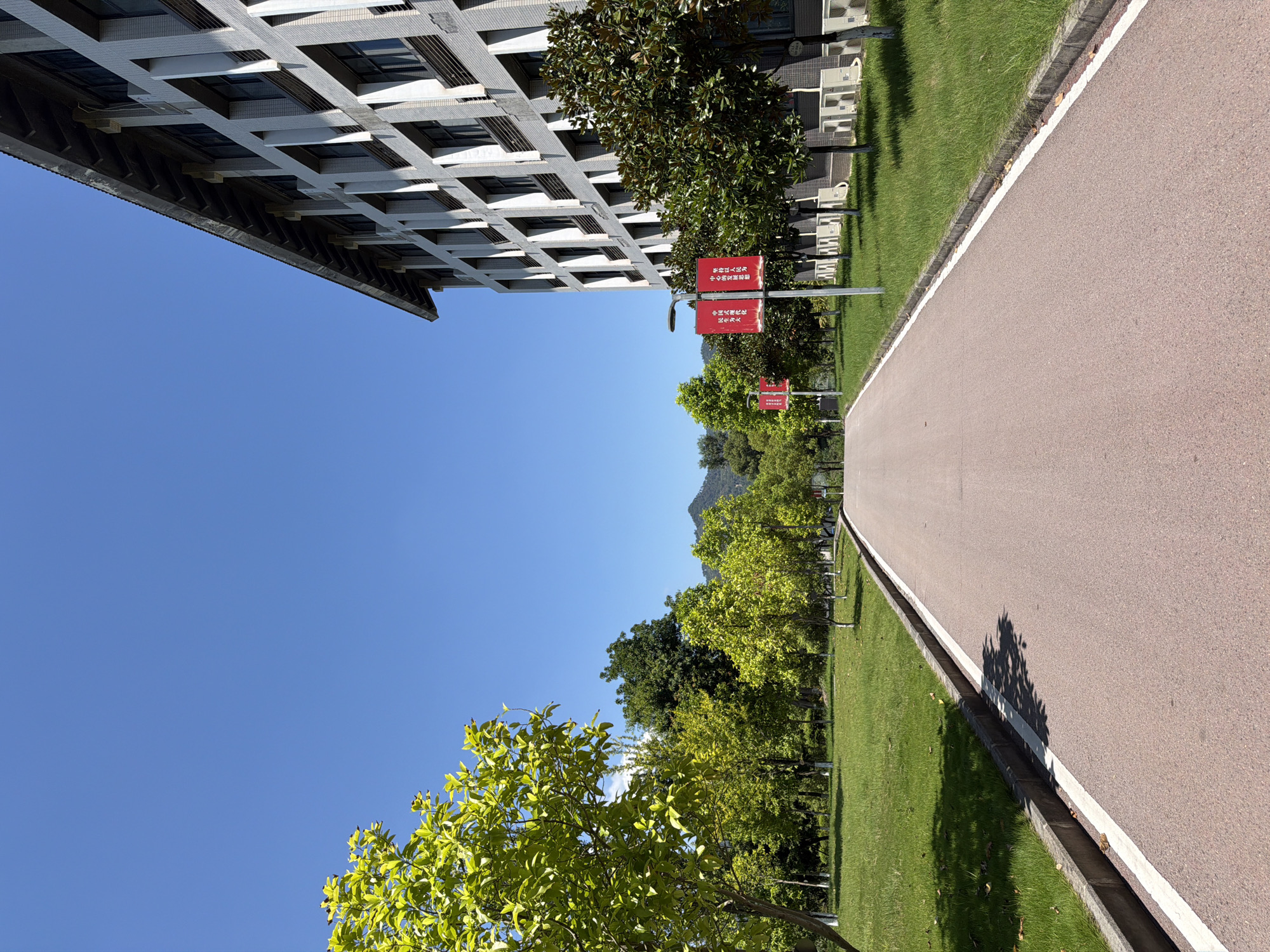 The image captures a view of a road leading into a campus setting, likely in the city of Jiangjin, China, given the context. The road is paved with a reddish-brown material and has white lines marking its edges. On the right side of the road, a large, modern building with a grid of windows and light-colored cladding dominates the frame. The building appears to be several stories high, with a distinctly sloped roofline. In front of the building, there is a neatly manicured lawn and several trees with dark green foliage. Red banners with Chinese characters are displayed on poles along the side of the road, suggesting an event or informational display. On the left side of the road, there is a grassy area with several trees, some with bright green foliage and others with darker leaves. The trees are spaced out, creating a park-like atmosphere. In the background, there are more trees and rolling hills or mountains under a clear blue sky. The lighting suggests it is a sunny day, with shadows cast by the trees onto the grass and road. There are no people visible in the image. The overall impression is one of an orderly and well-maintained institutional or educational environment.