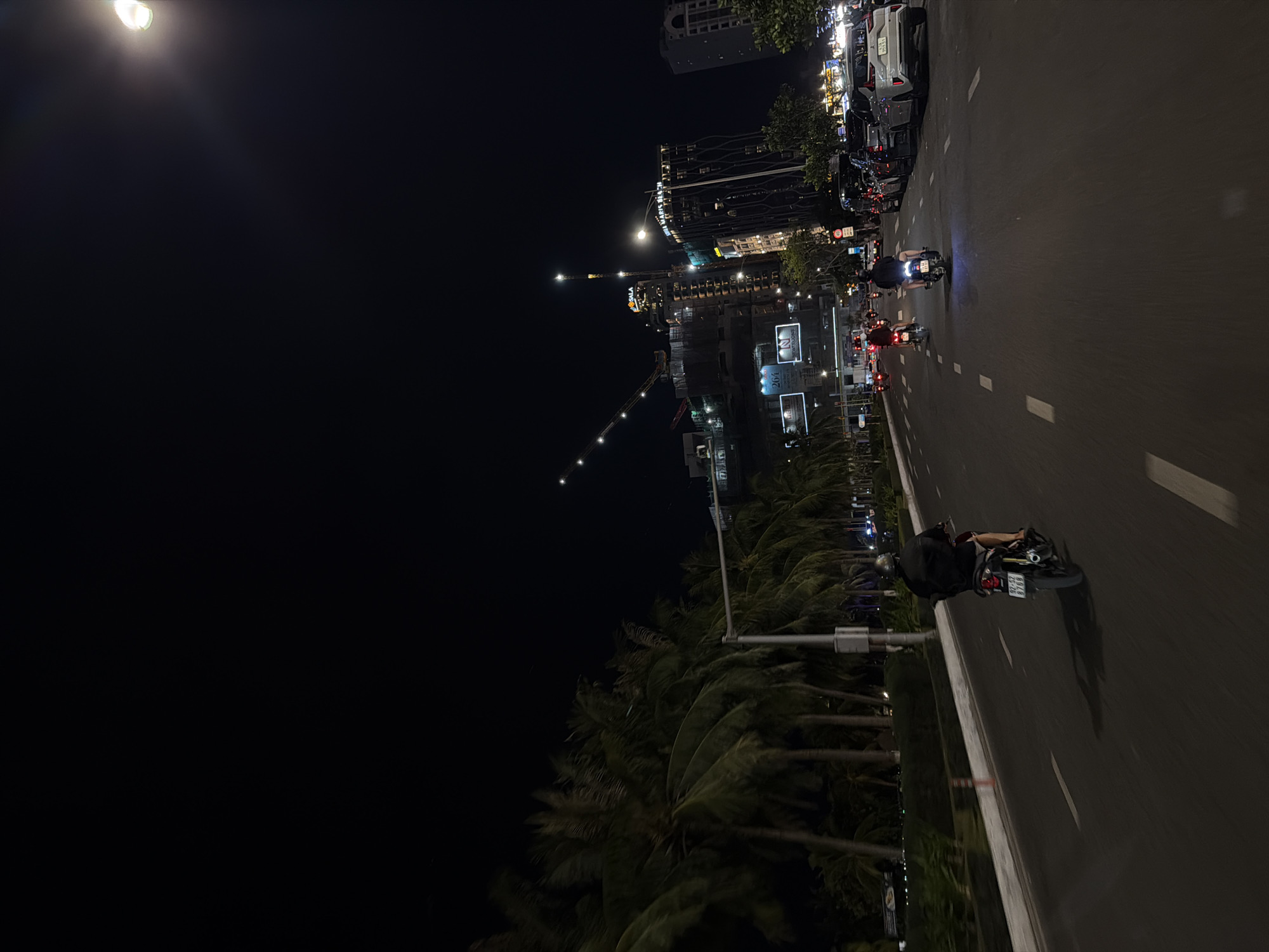 This image captures a nighttime scene on a busy street in Da Nang, Vietnam, likely near the My Khe Beach area, given the presence of palm trees lining the road and the visible construction of high-rise buildings in the background.
The foreground features a dark asphalt road with white lane markings. Two individuals on motorcycles are traveling away from the camera. The motorcycle on the left is closer and its rider is wearing a dark jacket and shorts. The motorcycle on the right has its headlights illuminated. Several cars are parked along the right side of the road.
In the mid-ground, a row of palm trees is visible on the left, their fronds creating a natural barrier. A light pole stands next to the road. Further back, several tall buildings are under construction, illuminated by cranes and lights. One building prominently displays the "SALA" logo. Another building to its right is illuminated and appears to be a completed structure.
The sky is dark, with the bright light of a streetlamp visible in the upper right corner. The overall impression is of a vibrant city at night, with ongoing development and traffic.