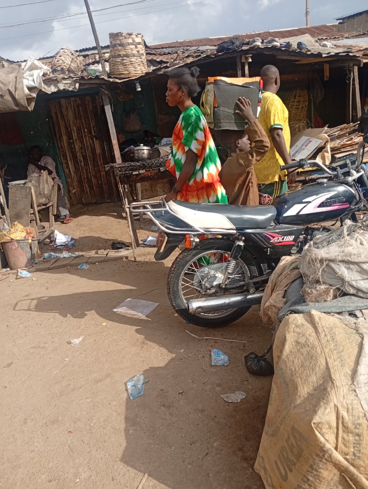 In Rafin Par, Nigeria, this image depicts a busy street scene near a marketplace. A woman in a tie-dye shirt walks past a stall with a thatched roof. She is followed by a boy carrying a large bag on his head and a man in a yellow shirt. A motorcycle is parked nearby, partially obscured by large sacks. The ground is unpaved and littered with debris, including plastic bags and paper. The stalls appear to be made of corrugated metal and wood, suggesting a temporary or informal market setting. The overall impression is one of commerce and daily life in a Nigerian urban environment.