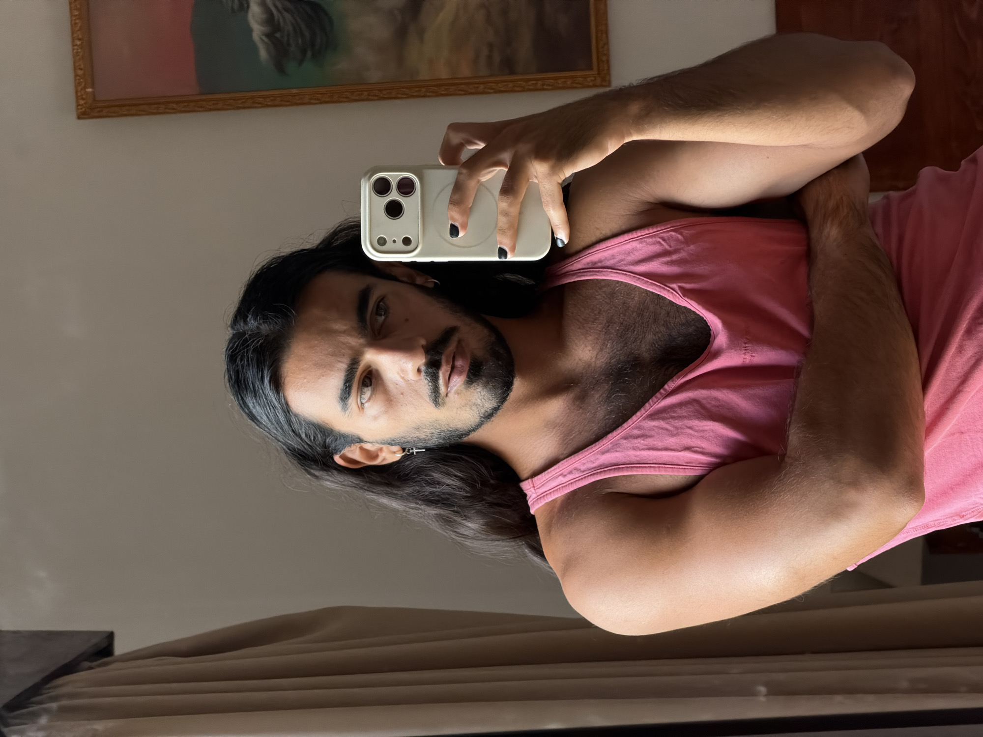 A man with long dark hair, a mustache, and a beard is taking a selfie in a room. He is wearing a pink tank top and has a silver cross earring in his left ear. His right arm is crossed over his chest. He is holding a white smartphone in his right hand, which is facing the camera. The background shows a window with a beige curtain on the left and a framed painting on the right. The image is a portrait taken indoors, with natural light illuminating the subject. The location is not identifiable within the city of North Kuta, Indonesia from the visual information provided.