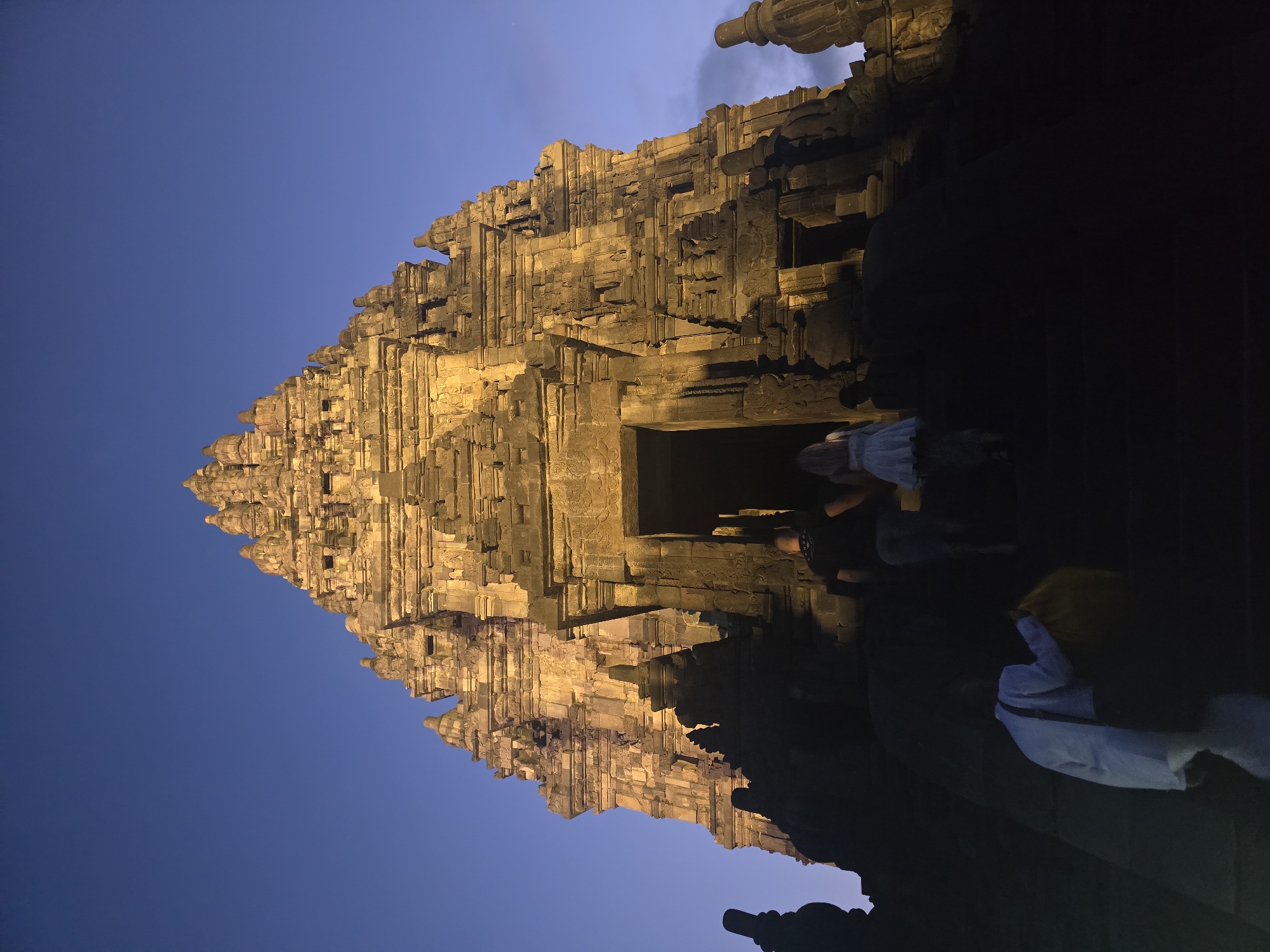 Captured at night in Bokoharjo, Indonesia, the image depicts a multi-tiered stone structure, illuminated in a warm golden glow against a twilight sky. The structure, likely a temple, is built of stacked stone blocks, with intricate details and carvings visible on its surface. The lower portion of the image is obscured by shadow, with several figures visible on the right side, including individuals with light-colored clothing.