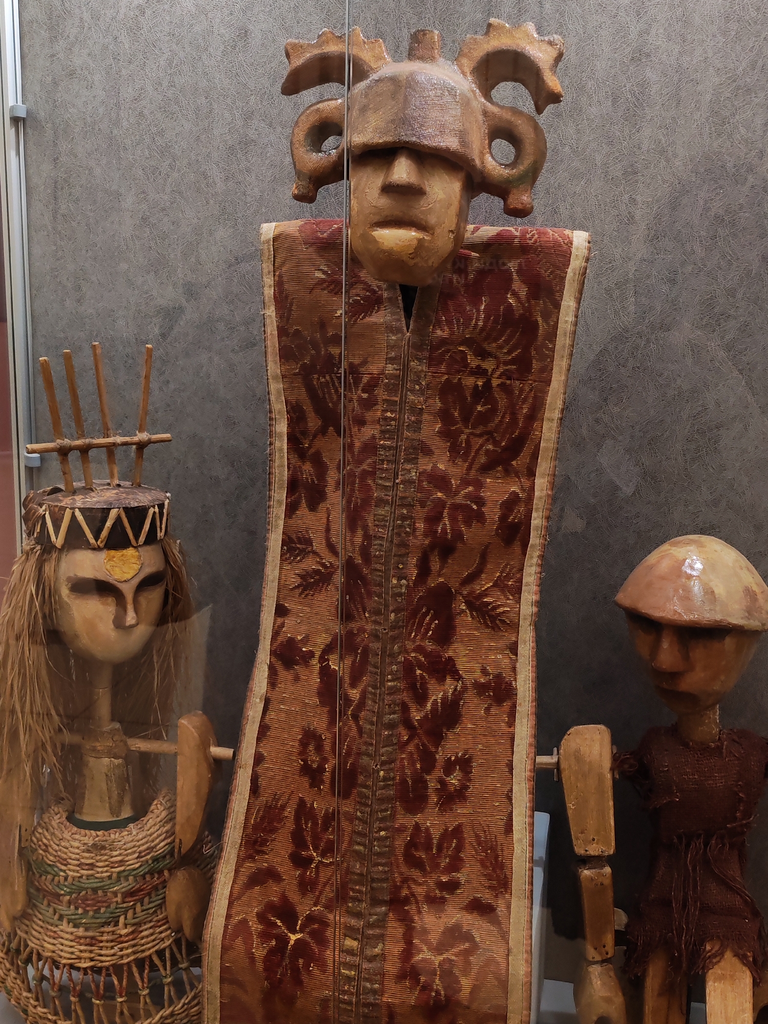 The image shows a display case, likely in a museum. Three figures are visible, probably artifacts. The central figure is tall, adorned with a reddish-brown, patterned textile. It features a sculpted head with a helmet-like headpiece decorated with curled elements. To the left, a figure with a straw-like headdress and woven dress is present. A smaller figure with a simple, dome-shaped headpiece and tattered garment stands on the right. The display case appears to be located in Izhevsk, Russia, though this location is not visually verified in the image.