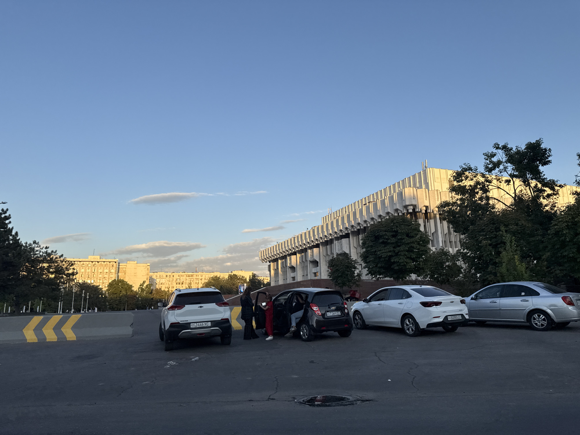 A street scene in Tashkent, Uzbekistan, features a modern building with a distinctive patterned facade bathed in warm sunlight. Several cars are parked in a lot in front of the building. Among them is a white Chevrolet Tracker with license plate 01 J446 XC. A black Chevrolet Spark, with license plate 01 TD69 ZC, has its doors open and two people are standing beside it, appearing to be interacting. A white Chevrolet Nexia is parked to the right of the Spark, followed by a silver Chevrolet Lacetti. In the background, apartment buildings are visible, and the sky is a clear blue with a few wispy clouds. The ground is asphalt, with visible cracks and a manhole cover. Yellow and black striped concrete barriers are positioned on the left side of the frame. The overall impression is a snapshot of urban life during daylight hours.