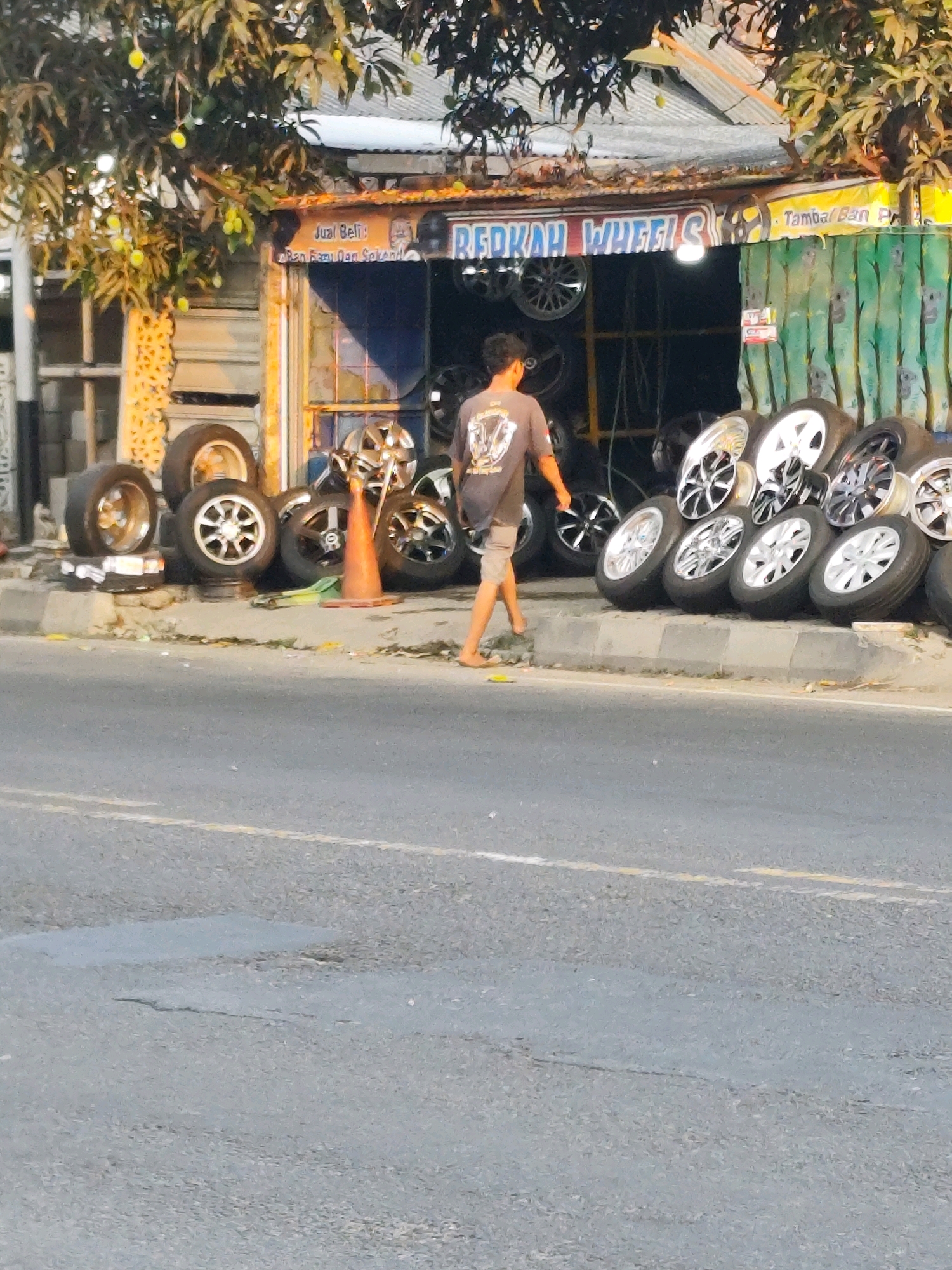 In the city of Bantul, Indonesia, a man is walking past a tire shop on a street. The shop, named "REDCAH WHEELS," displays a variety of tires and rims. The man, wearing a t-shirt and shorts, is walking on the sidewalk towards the right of the frame. Trees with fruit are visible in the background. The street in front of the shop has some cracks and appears to be a road.