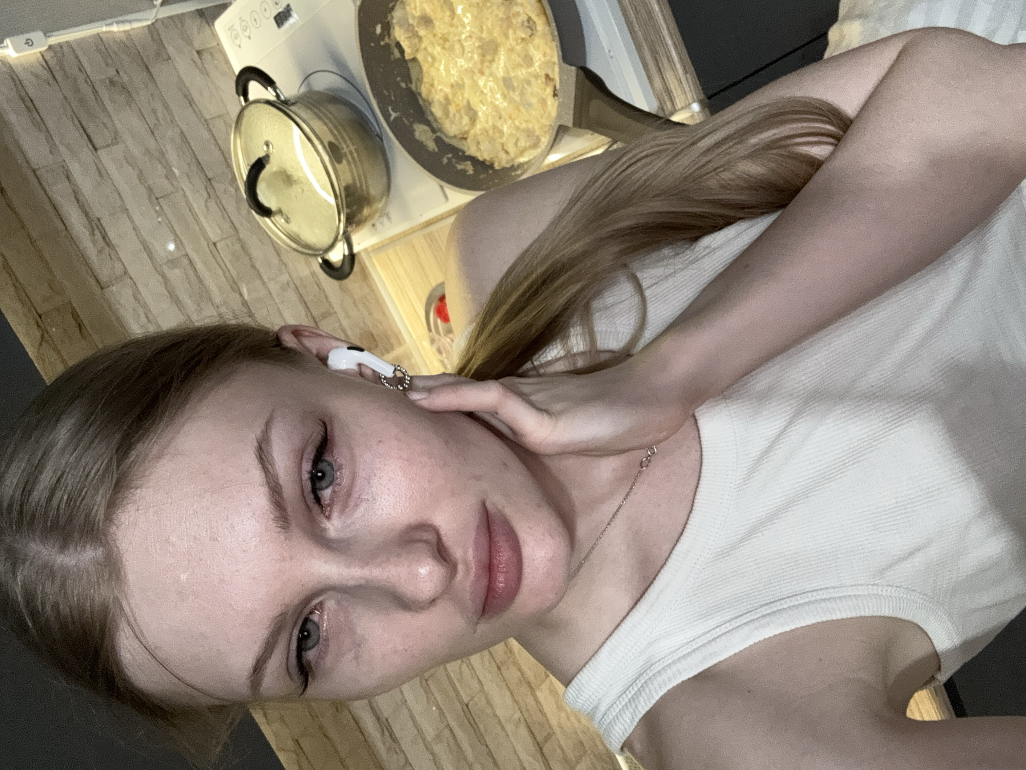 A selfie of a fair-skinned woman with blonde hair, wearing a white tank top, is positioned in front of a kitchen scene. A pot with a lid and a frying pan filled with a creamy, cooked dish rest on top of a stove. The background features a tiled wall with a light-colored, possibly beige, wood-grain pattern. The woman has one hand touching her neck. The presence of a pot and pan with food suggests a domestic kitchen setting. There is no evidence in the image to pinpoint the location to Perm, Russia.