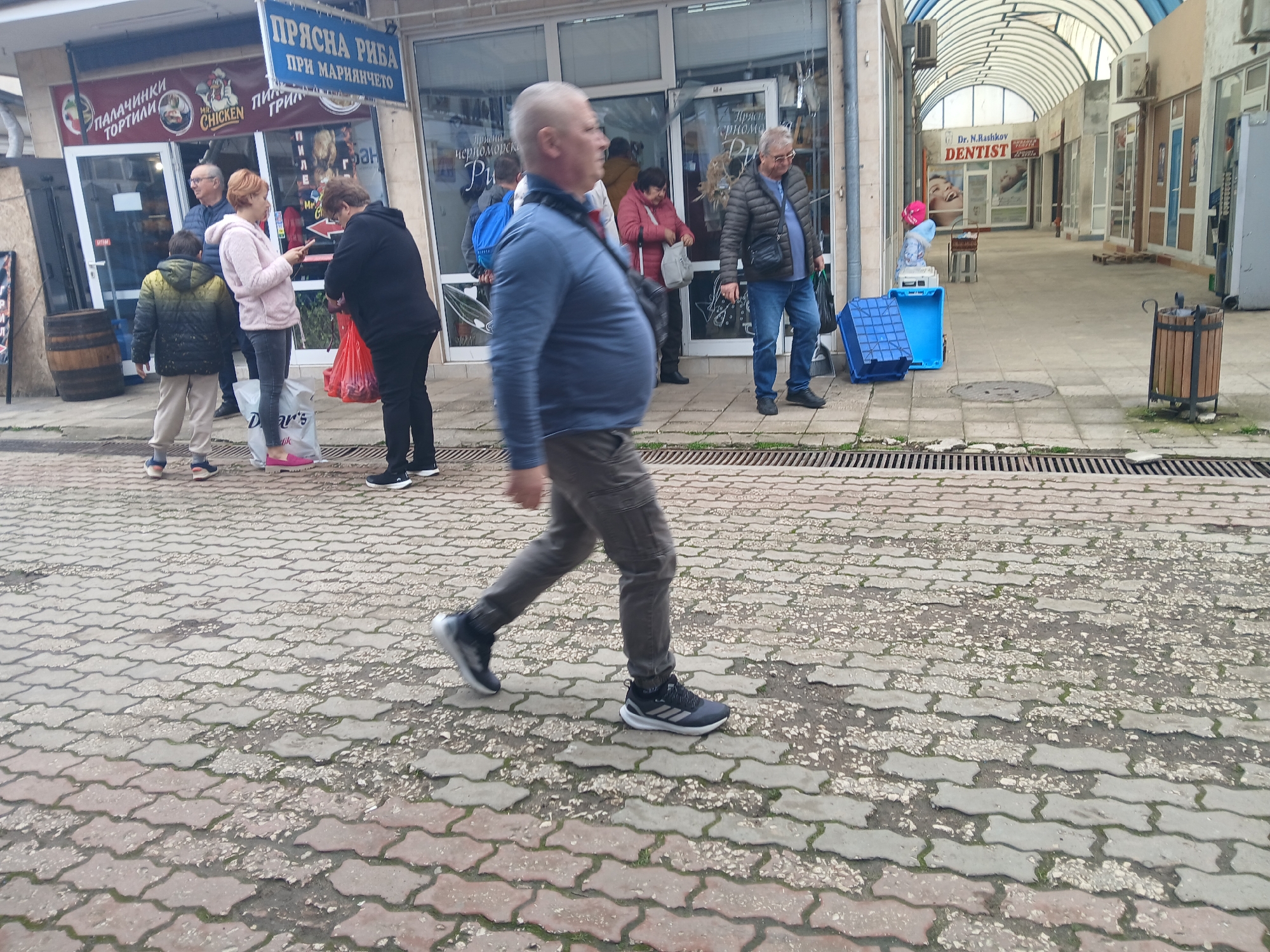 This medium, wide shot captures a group of people walking in Kavarna, Bulgaria. In the foreground, an older, bald man walks left, wearing a blue shirt, gray pants, and black sneakers with white soles. Behind him, several people stand near a storefront advertising "Fresh Fish at Mariyancheto," along with "Mr. Chicken Grill" and "Pancakes." These people are looking at their phone and shopping.
In the mid-ground to the right, an older man in a quilted jacket and jeans stands near a stack of blue plastic crates. Further down the covered pedestrian walkway is the office of Dr. N. Rashkov, Dentist. The ground is paved with light-colored interlocking pavers, some of which show signs of wear. A small trash can is visible to the right side of the photo. The lighting suggests an overcast day.