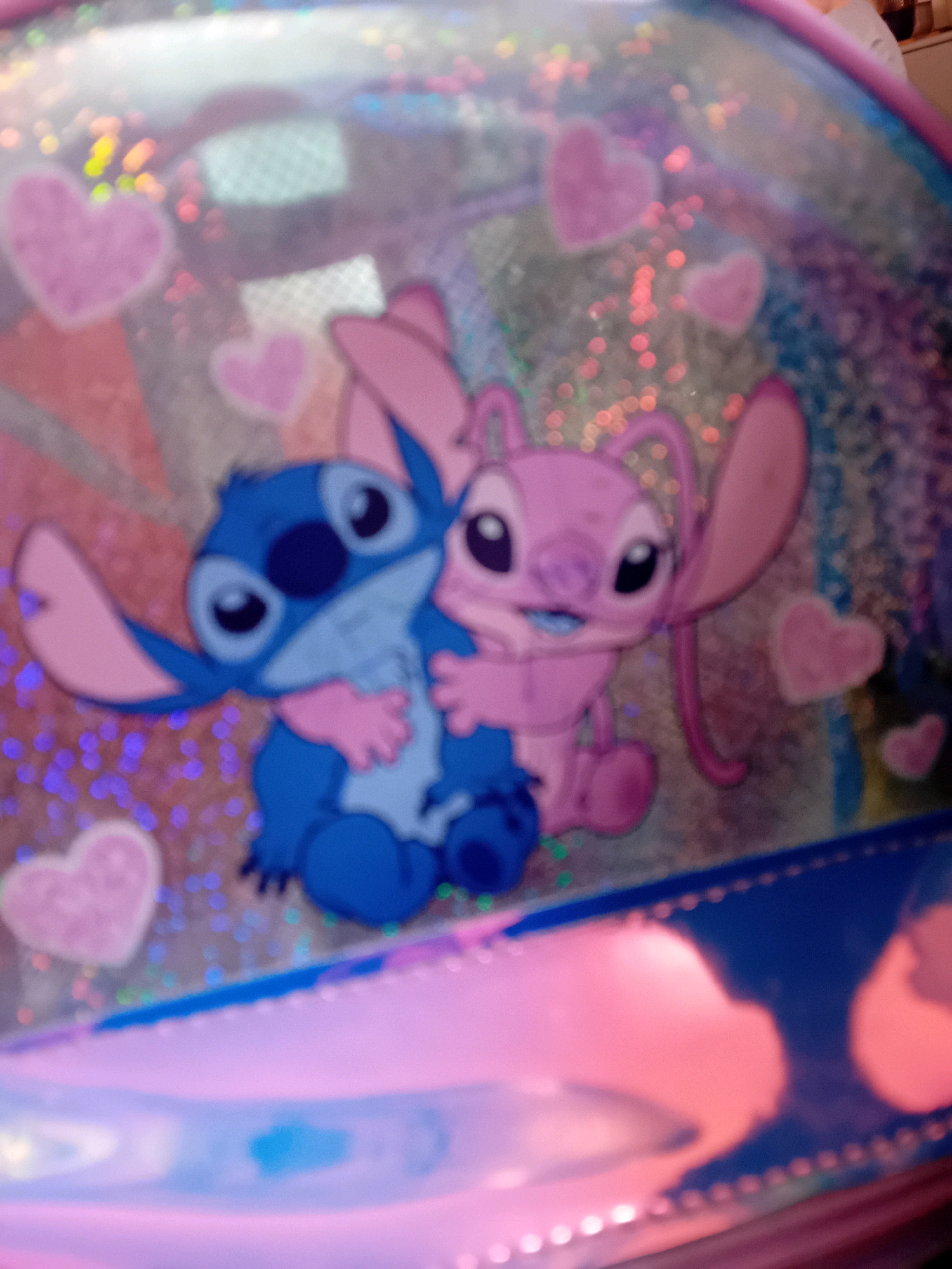 The image shows a close-up of a product, likely a backpack or a bag, featuring cartoon characters Stitch and Angel from Disney's Lilo & Stitch. They are depicted embracing each other, with Stitch on the left and Angel on the right. The background is sparkly and iridescent, with pink hearts and bokeh effects, suggesting a whimsical or playful design. The product itself appears to be made of a clear, shiny material over a pink and blue base. There are no people, specific locations, or any indication of the city of Chuhuiv, Ukraine, within the image. The focus is entirely on the characters and the product's design elements.