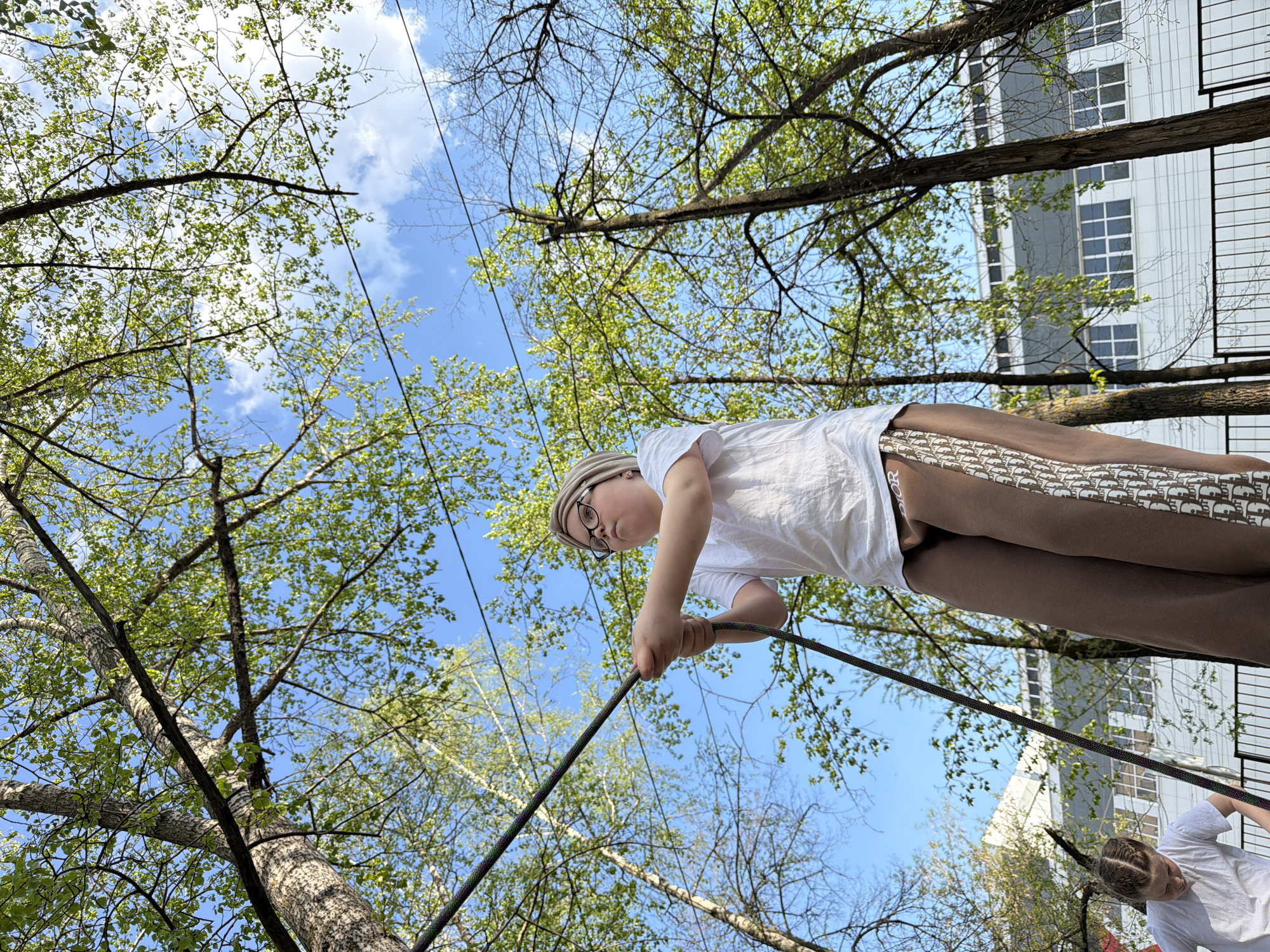 The image shows a low-angle view in what appears to be a park or green space. A child with glasses and a light-colored cap is holding onto a dark rope extending from the bottom center of the frame upwards through tree branches. The child is wearing a white t-shirt and brown pants with a repeating pattern. The sky is blue with some clouds visible through the tree canopy. Tree branches with green leaves fill much of the frame. In the background, the facade of a building with a grid of windows is visible. Another child's arm and torso are also visible towards the bottom-right of the image. The photo was taken in Izhevsk, Russia.