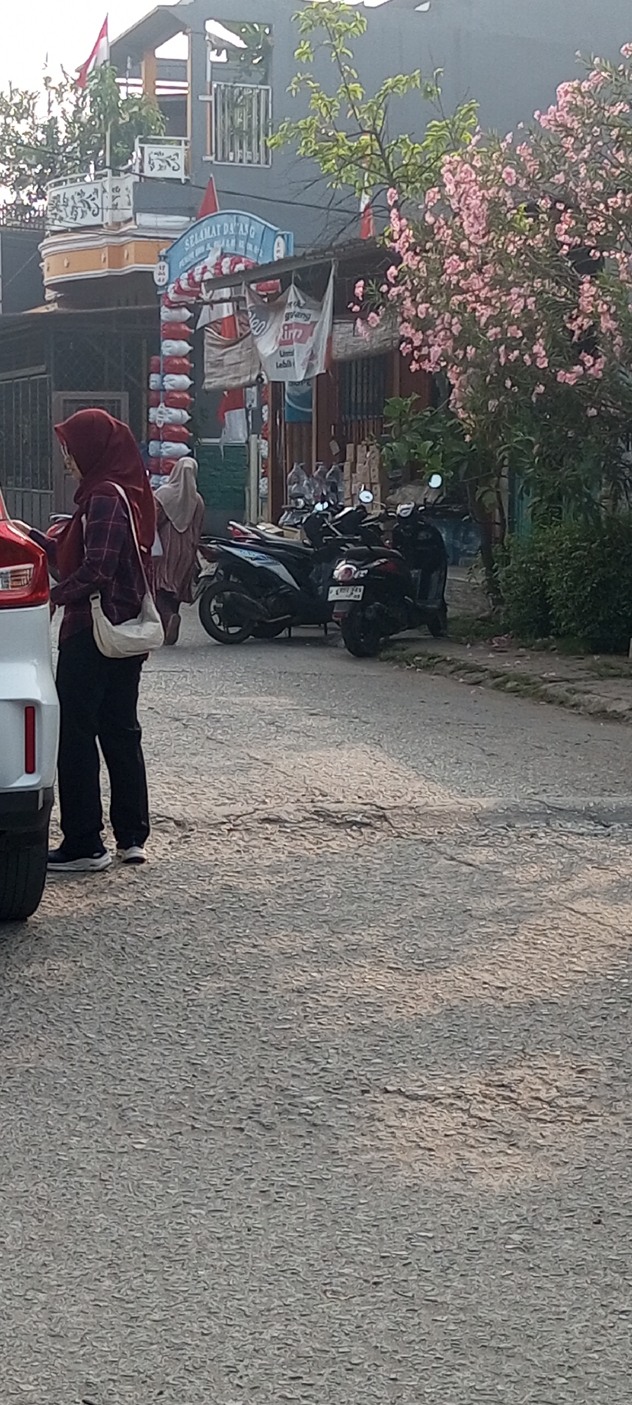 A side view of a person wearing a maroon hijab, a plaid shirt, black pants, and white shoes stands near a white car on a street in Setu, Indonesia. The person is looking at their phone. In the background, several motorcycles are parked on the street. A building with a blue sign that reads "SELAMAT DATANG" (Welcome) is visible. Pink flowers bloom on a tree to the right of the frame. The street surface is cracked and uneven.