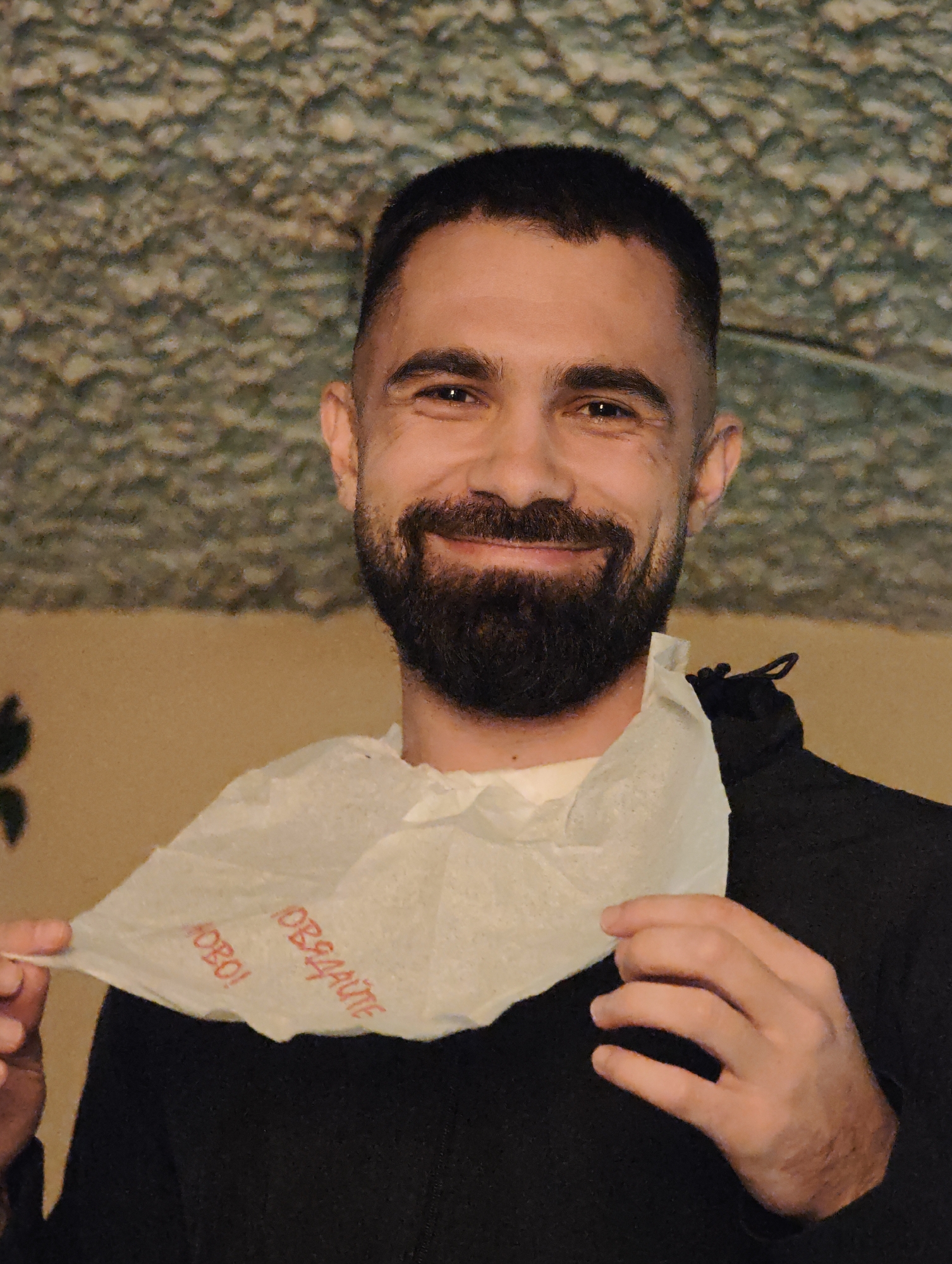A man with dark hair and a beard smiles while holding a white bib with red Cyrillic text. The text appears to read "Hobo! Обдайте." He wears a black hooded jacket. The background consists of a mottled wall texture, and the lighting is ambient. This is likely an indoor setting, possibly a restaurant or home. The city of Sofia, Bulgaria is mentioned in the prompt, but there is no other information to ascertain if that is the correct location.