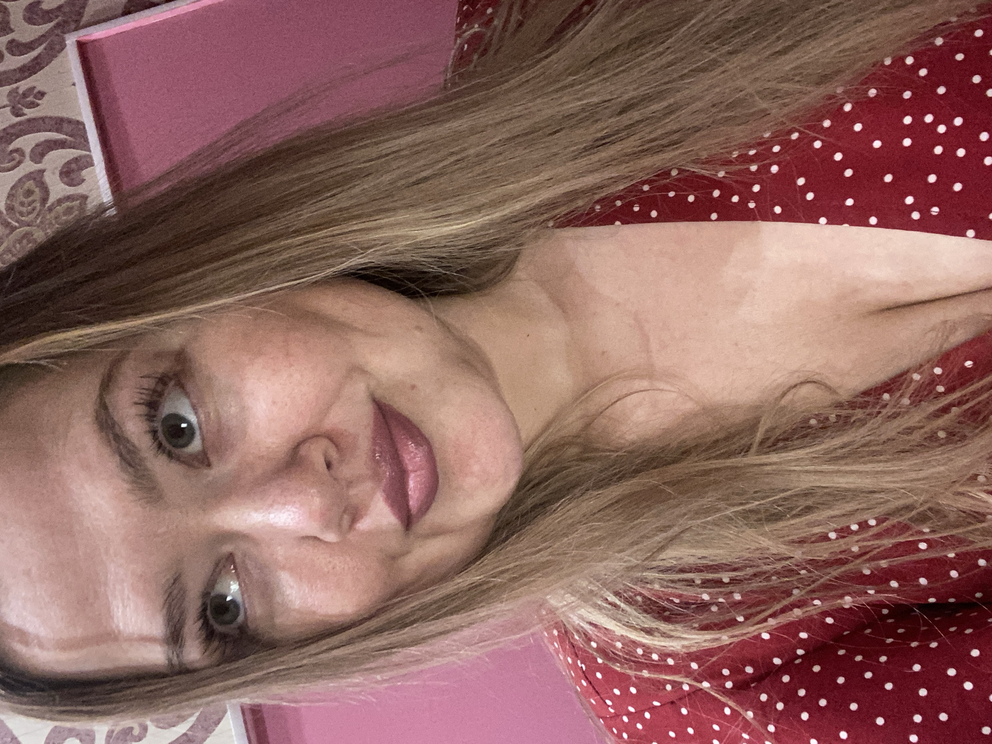 A blonde woman with fair skin and light blue eyes poses for a selfie. She is wearing a red dress with white polka dots. The background appears to be a wall with pink and patterned wallpaper. The image does not provide any information about the location being in Yerevan, Armenia.