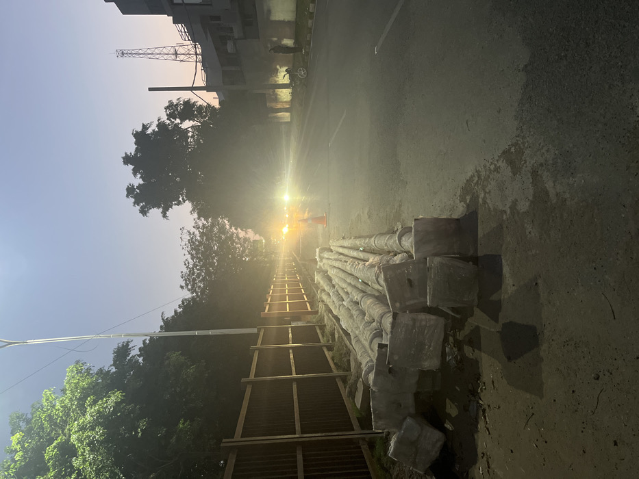 The image shows a stack of concrete utility poles lying on the side of a paved road in Sahibzada Ajit Singh Nagar, India. The road appears empty, and there is a light mist or haze in the air. There is a wooden fence and trees lining the side of the road. A streetlight is visible in the background, and there is a small orange traffic cone near the stack of poles. The scene is lit by the streetlights and the headlights of an approaching vehicle. The time of day appears to be evening or early morning.