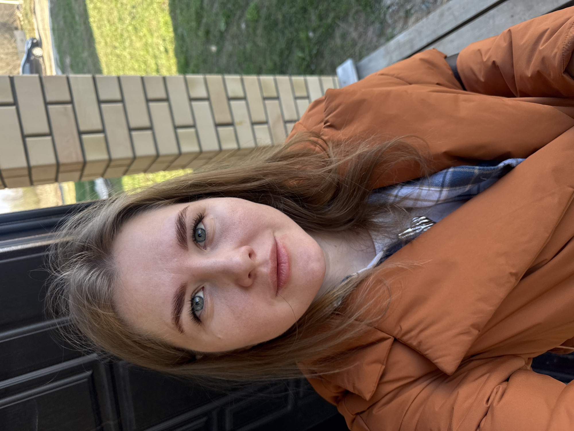 A young woman is centered in the frame, wearing an orange puffer jacket and a blue and white plaid shirt underneath. She has fair skin, light brown hair, and blue eyes. She is positioned in front of a dark, possibly black, paneled surface. Behind her, a brick wall and a grassy area are visible, suggesting an outdoor location. The photo is taken in the city of Ферма, Russia.