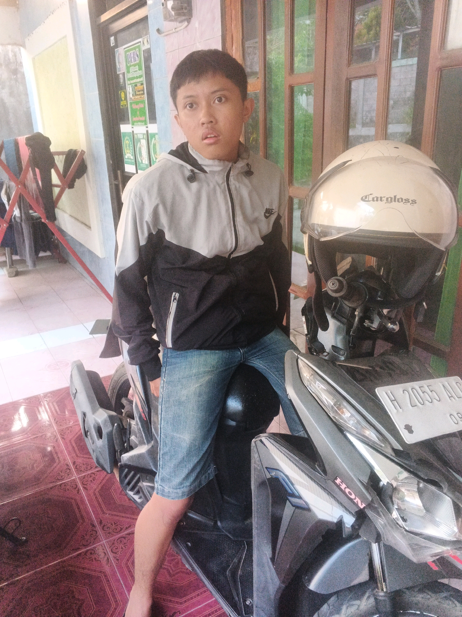 A young East Asian male sits on a dark gray motorcycle in front of a building with wooden framed windows and doors. He is wearing a light gray and black jacket and blue denim jeans. A white helmet with "Cargloss" written on it is resting on the motorcycle's handlebars. A license plate on the motorcycle reads "H 2055 ALC 09". In the background, clothing hangs on a drying rack. The scene appears to be outdoors, possibly in a residential area of Keji, Indonesia, indicated by the architecture and common motorcycle usage.