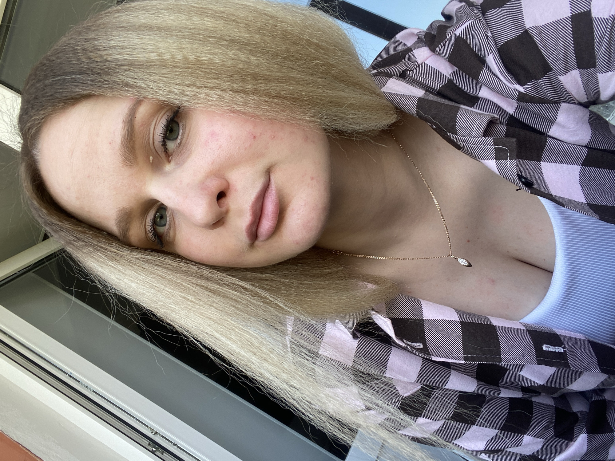 An eye-level selfie captures a young woman inside a building in Chaikovsky, Russia, looking directly at the camera with a slight head tilt. She has long, blonde hair with a textured, almost crimped appearance, parted on the side and falling across her shoulders. Her eyes are light-colored, possibly green or hazel. Her face shows natural skin texture, including visible pores and some minor redness or blemishes on her cheeks and forehead. A small, white bump is visible beneath her left eye. She wears a button-up shirt with a pink or light purple and black or dark gray plaid pattern, worn over a white, ribbed tank top or camisole. A delicate gold chain necklace with a small, leaf-shaped clear pendant rests on her chest. The background features a blurred white and dark-colored window or door frame, suggesting an indoor setting with bright natural light coming from outside, indicating it is daytime. Her expression is neutral to softly contemplative, with a faint hint of a smile. No text is visible in the image.