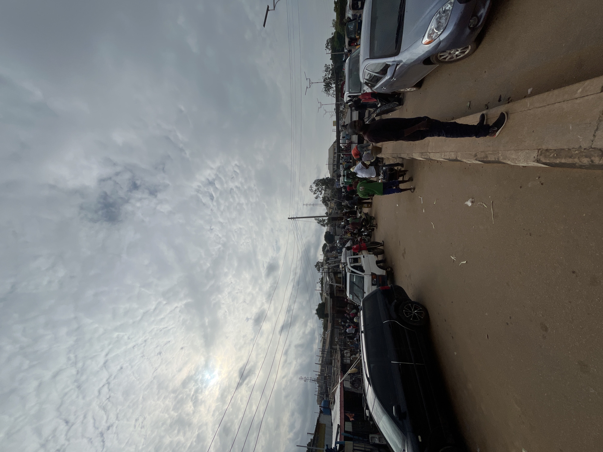 The image captures a street scene in Gura Top, Nigeria, under an overcast sky. The perspective is rotated, with the road and sidewalk running vertically. A line of vehicles, including a dark SUV in the foreground and a silver car and white van further up the street, are parked or moving along the road. Several people are visible on the sidewalk and in the street, some standing in groups, others appearing to be engaged in commerce or simply passing by. The overall impression is of a busy, everyday street life. The weather appears to be cloudy, with the sun faintly visible through breaks in the clouds, suggesting it might be daytime, perhaps late morning or afternoon. There are no visible signs of text in the image.