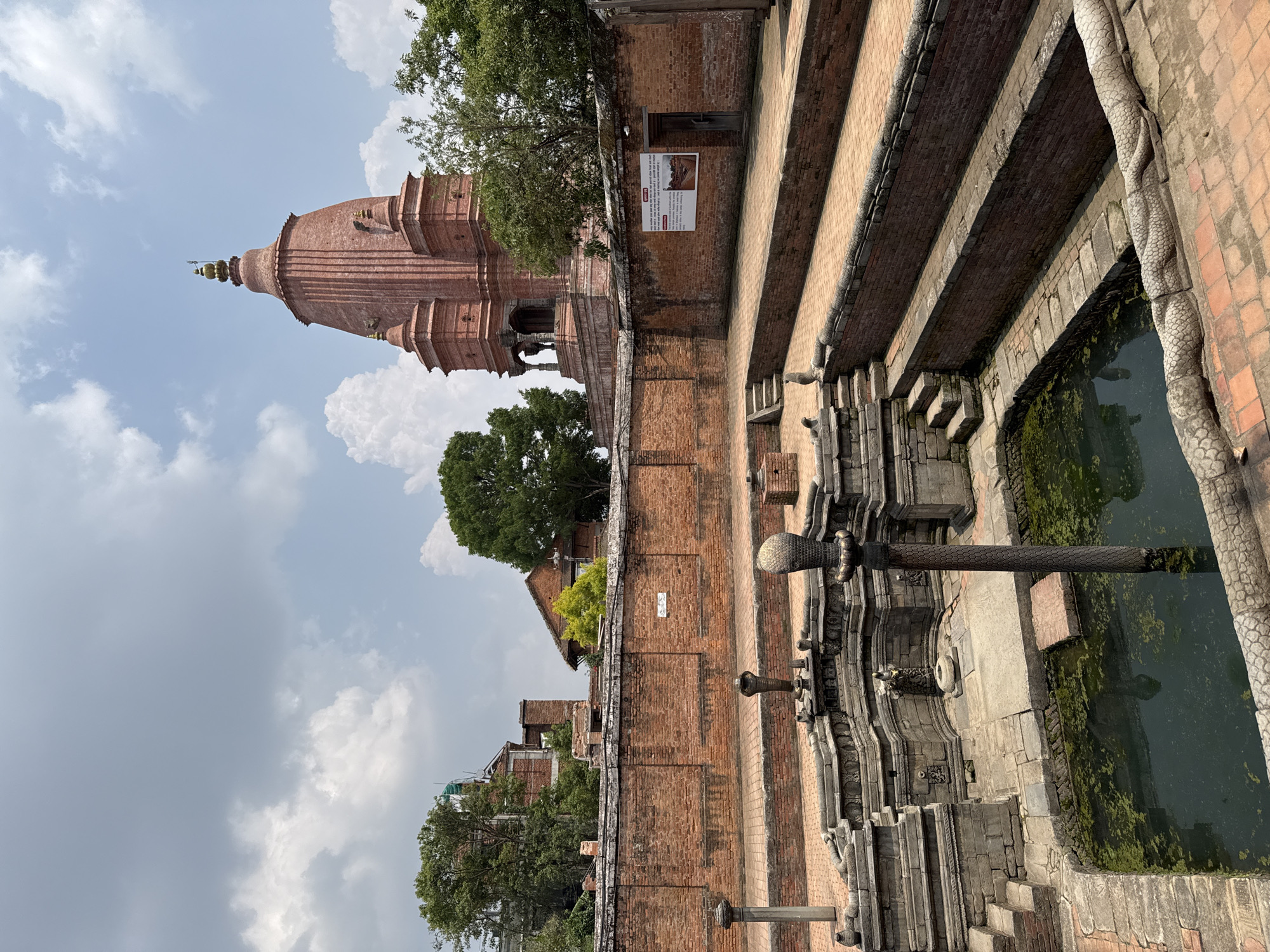 The image shows a view in Bhaktapur, Nepal, featuring the 55 Window Palace and a historic water tank. On the left, the red brick facade of the 55 Window Palace rises, adorned with intricate architectural details and topped with a towering spire. Adjacent to the palace, a stepped brick structure leads down to a water tank. The tank's edges are lined with stone carvings. The water appears murky, with greenish growth. A pipe extends horizontally above the water surface, positioned as a spout for water. The sky above is partly cloudy. The overall scene is one of historical architecture.