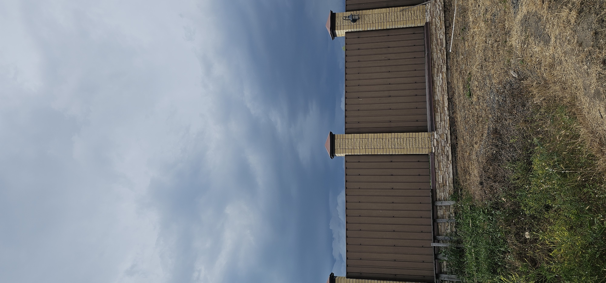 The media file displays a brown metal fence with decorative brick pillars topped with dark caps. A light fixture is visible on one of the pillars. The fence is bordered by a stone-clad base. To the right of the fence, there is dry, tall grass and some green vegetation. The sky above is a mix of blue and white clouds, suggesting daytime with potentially changing weather. The scene appears to be outdoors, possibly in a rural or suburban setting, given the vegetation and fencing. There are no people visible in the image. The overall impression is one of an exterior view of a property boundary. The location context indicates the scene is in Овощной, Russia.