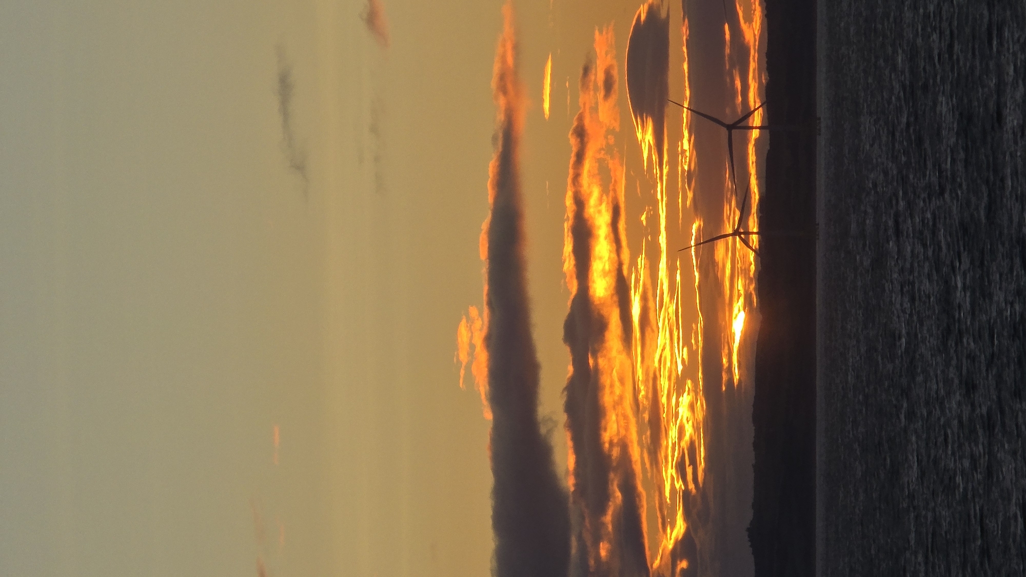 The image shows a sunset over a body of water, likely the sea, with a silhouetted coastline in the distance. Two wind turbines are visible in the foreground, positioned on the water, with their blades partially obscured by the sunset. The sky is a vibrant gradient of orange, yellow, and pale blue, with dramatic clouds catching the warm light. The water is dark and choppy, reflecting the colors of the sky. The overall impression is one of a serene and powerful natural landscape, with human-made structures integrated into the environment. The specific location in the United Kingdom is not identifiable from the image.
