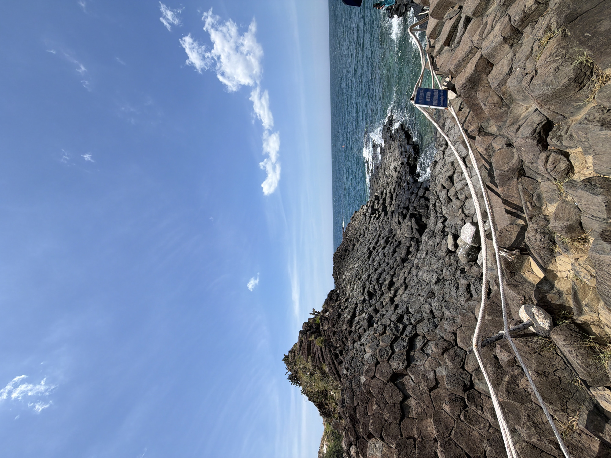 The image depicts a scenic view of the basalt columns of Gành Đá Đĩa in the commune of An Ninh Đông, district of Tuy An, in the city of Phú Lương, Vietnam. The foreground is dominated by a rugged expanse of dark, hexagonal basalt rock formations, forming a natural staircase-like pattern. A thick white rope, acting as a safety barrier, is strung across several wooden posts, guiding visitors through the rocky terrain. Beyond the rocks, a turquoise ocean stretches to the horizon, with gentle waves lapping against the shore, creating white foam. The sky is a vibrant blue, with scattered white clouds adding depth and texture to the scene. A small blue sign with white lettering is visible near the rope barrier, likely providing information or safety instructions. The overall atmosphere is one of natural beauty and geological wonder.