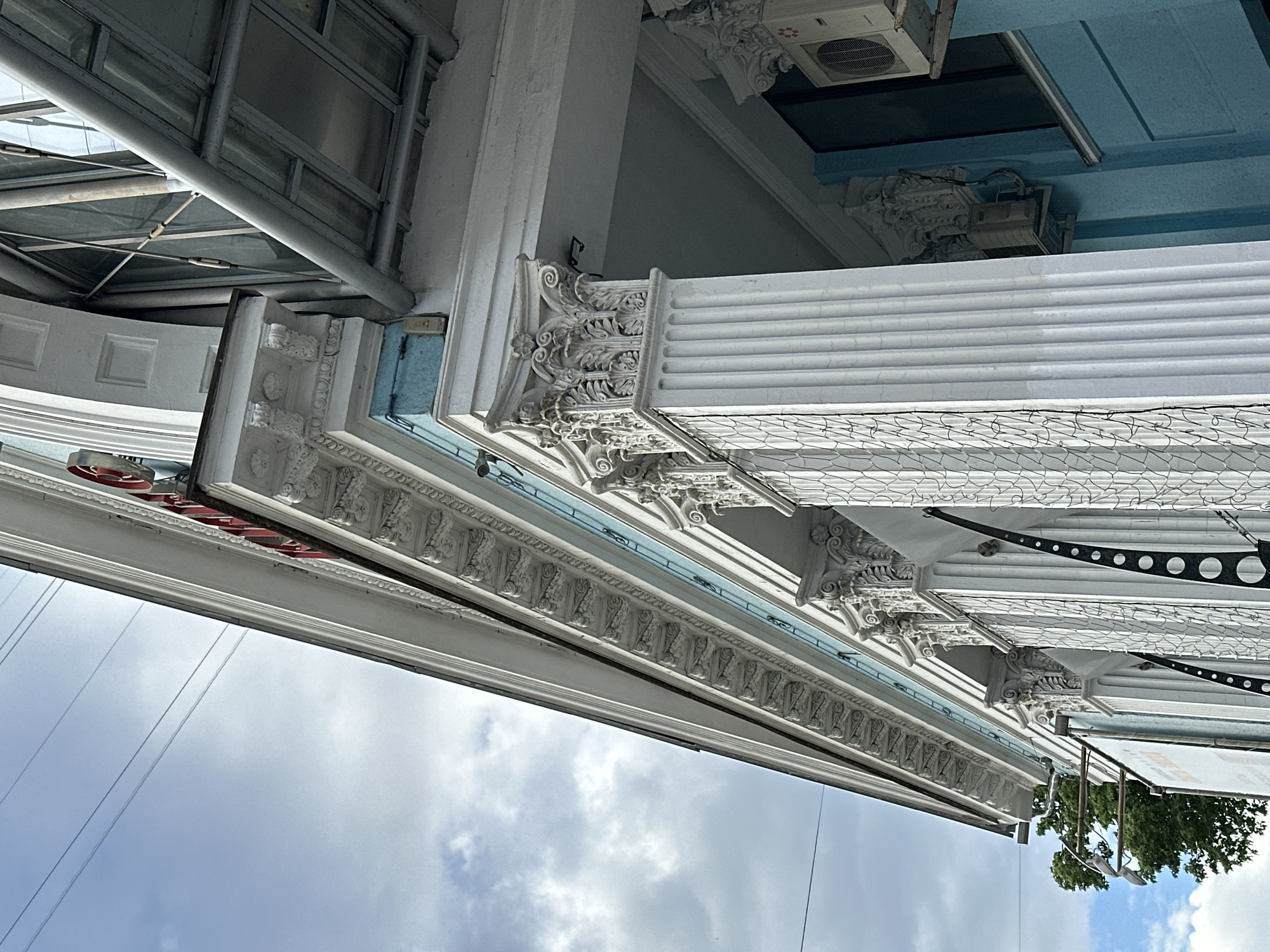 The image shows an upward-angled view of architectural details of a building in Moscow, Russia. The architecture features ornate white columns, decorative molding, and textured surfaces, which are partially obscured by modern infrastructural elements, like a metal awning or scaffolding. Above the facade, a sky with clouds and power lines is visible, while below and to the side are trees, suggesting a building close to the street. An air conditioning unit is affixed to the building's surface at the very top.
