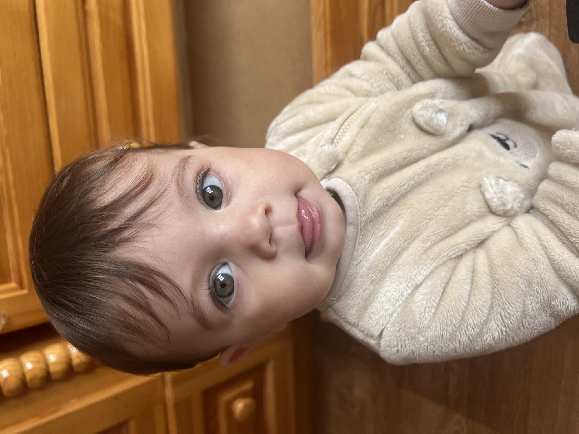A close-up, high-angle portrait shows a single infant with fair skin, dark brown hair, and large green-brown eyes looking directly at the camera. The infant's mouth is slightly open, with the tip of its tongue visible. The infant is wearing a light beige, plush, full-body sleeper featuring a central zipper and appliquéd elements resembling an animal, possibly a bear, on the torso. The background consists of blurred wooden furniture, specifically a dresser or cabinet with a warm reddish-brown finish, visible in the upper left and right, and a wooden floor in the lower left. The scene is indoors in Elena, Bulgaria.