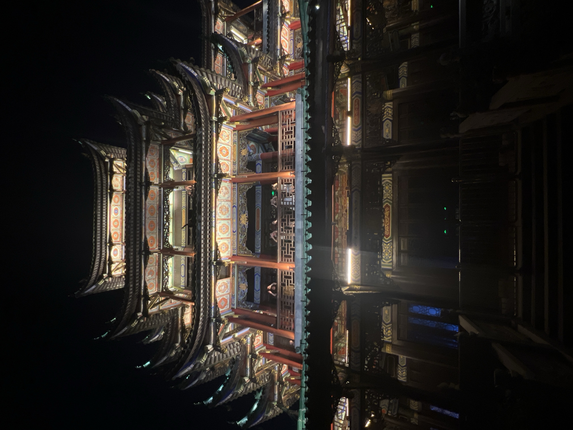 The image depicts a multi-tiered traditional Chinese building at night, illuminated from within, in the city of Liangjiang, China. The building features ornate, tiered roofs with upturned eaves, painted details in red, gold, and blue, and intricate wooden carvings. Several levels of balconies and walkways are visible, with railings and supporting pillars. A few silhouetted figures can be seen on one of the upper balconies, appearing to interact with each other. The foreground shows a dark staircase leading up to the building, with further darkened structures and railings flanking the stairs. The overall impression is of a grand, culturally significant structure set against a dark night sky.