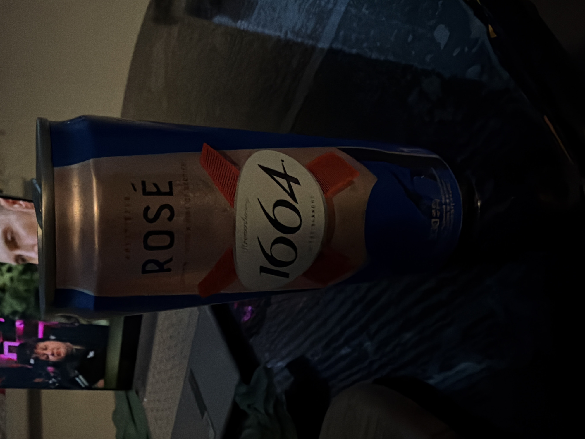 A close-up, dimly lit shot shows a can of Kronenbourg 1664 Blanc beer. The can has a blue and rose gold design. The text "ROSE" is prominently displayed on the can, along with the number "1664". In the background, a television screen displays an image of a woman. The scene is set indoors, likely on a table with a textured surface. The image appears to be taken in Bila Tserkva, Ukraine.