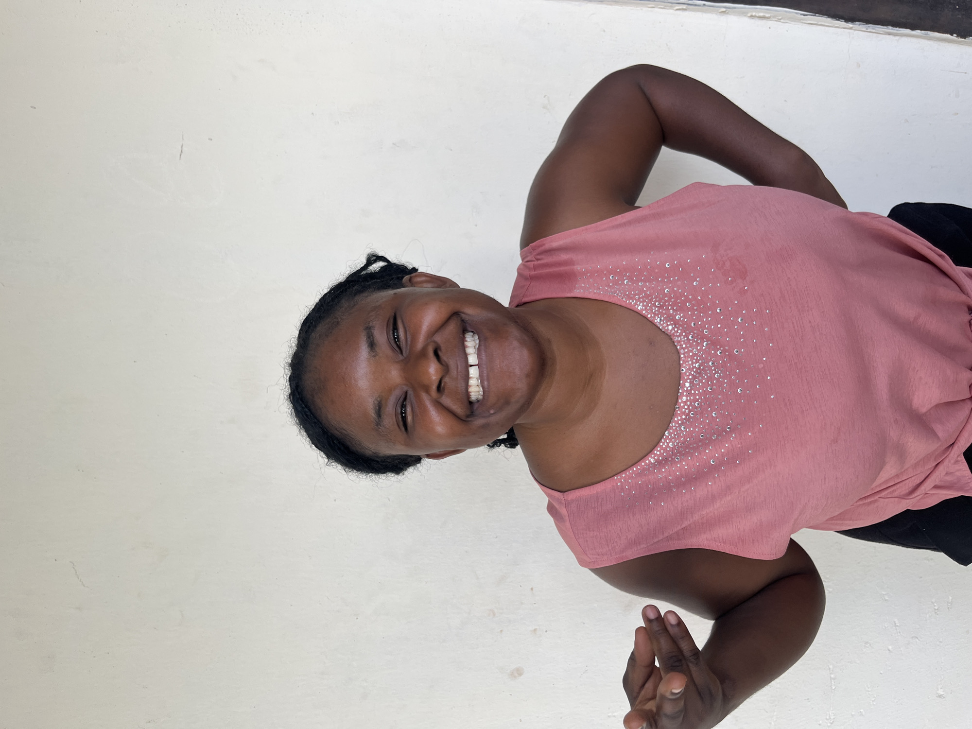 A woman is photographed from the chest up, turned to her right and looking at the camera with a wide, joyful smile. Her dark hair is braided and pulled back. She is wearing a light pink t-shirt adorned with a pattern of small, sparkling embellishments around the neckline. Her right arm is bent at the elbow, with her hand near her head. Her left hand is visible in the lower right corner of the frame, fingers slightly splayed. The background is a plain, off-white wall, possibly with some subtle discoloration or texture. The lighting appears to be from an overhead source, creating soft shadows. The image seems to capture a candid moment of happiness. The subject's clothing and the simple background suggest a casual, everyday setting. The image quality is clear, with good focus on the woman's face and upper body. There is no visible text in the image.