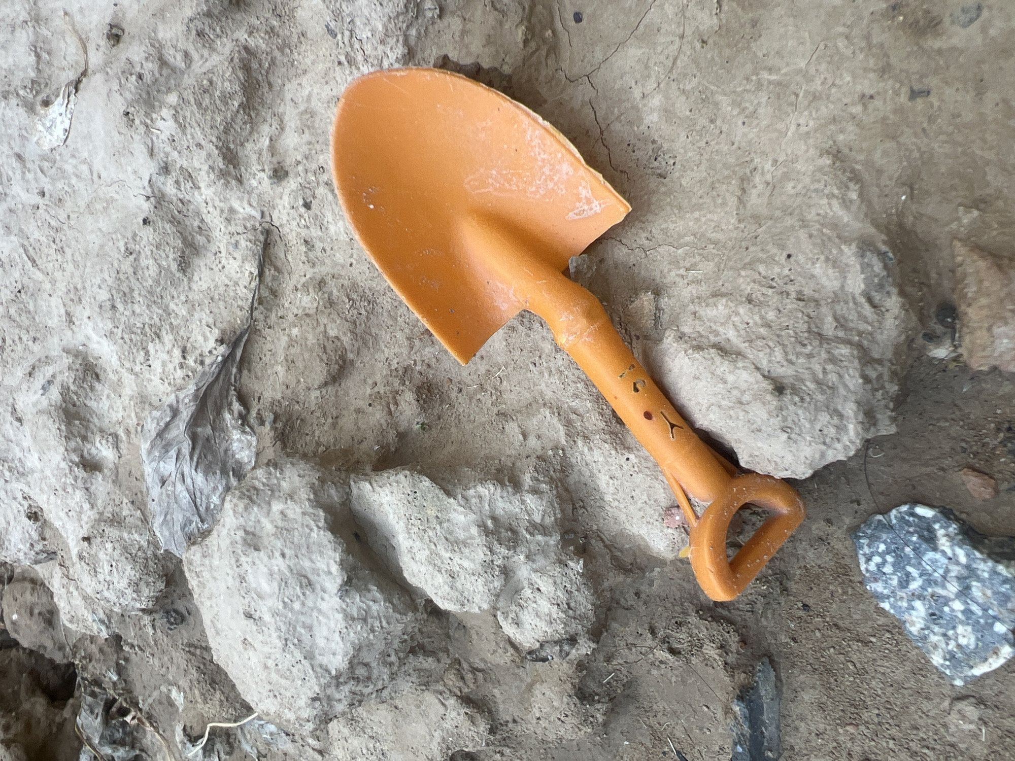 An orange toy shovel with a smiley face is resting on the ground among rocks and dirt. The shovel is positioned diagonally, with its handle pointing towards the bottom left and its blade towards the top right. The handle has several circular perforations and a smiley face appears to be drawn on it. The ground is a mix of light brown dirt and larger, light-colored rocks with rough textures. There are also some dried leaves and smaller pebbles scattered around. The lighting suggests it is daytime, with shadows indicating the direction of the light source. The image appears to be taken from a slightly elevated angle, looking down at the shovel and its surroundings.
